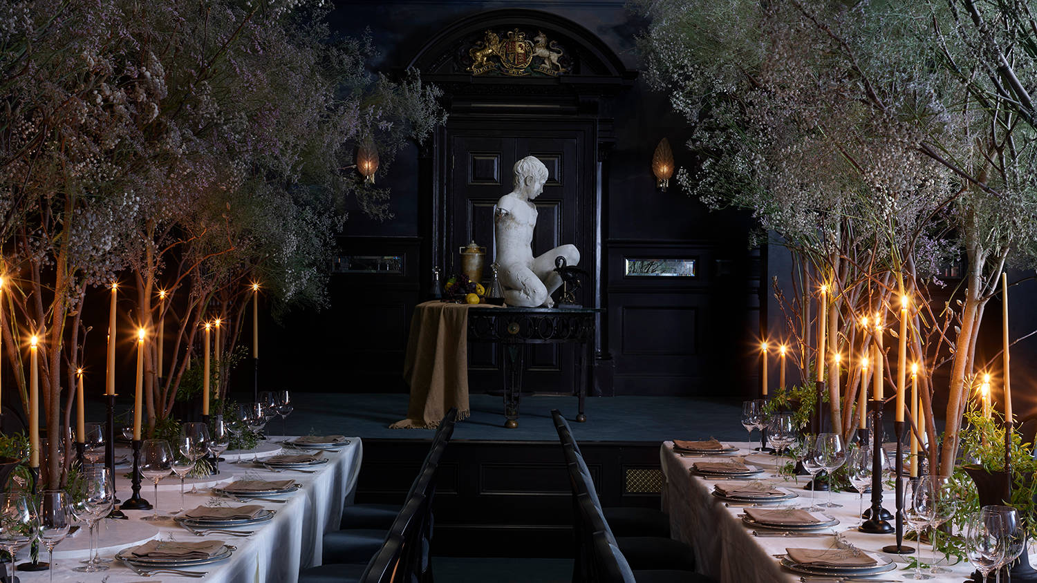 A grand dining room with two long tables, lit candles, tall branches, dark walls, and a stormy mural on the walls and ceiling.