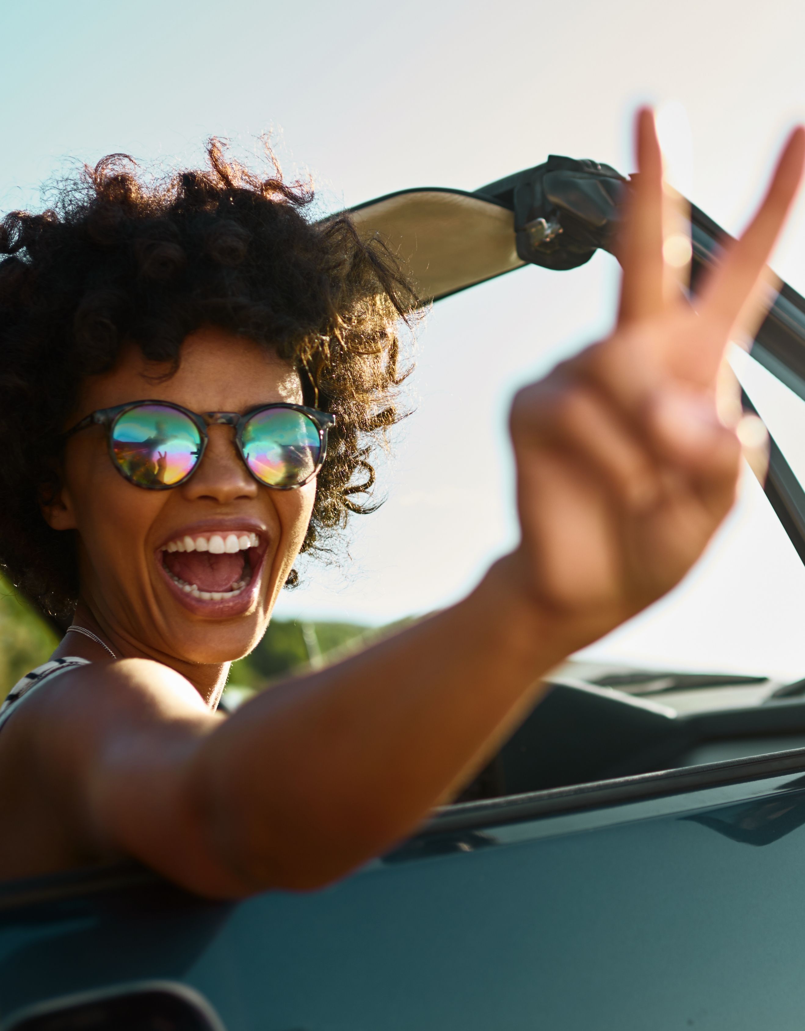 Young woman wearing a striped top and mirrored sun glasses sitting in a classic open top car looking into the camera making a  peace sign with her right hand