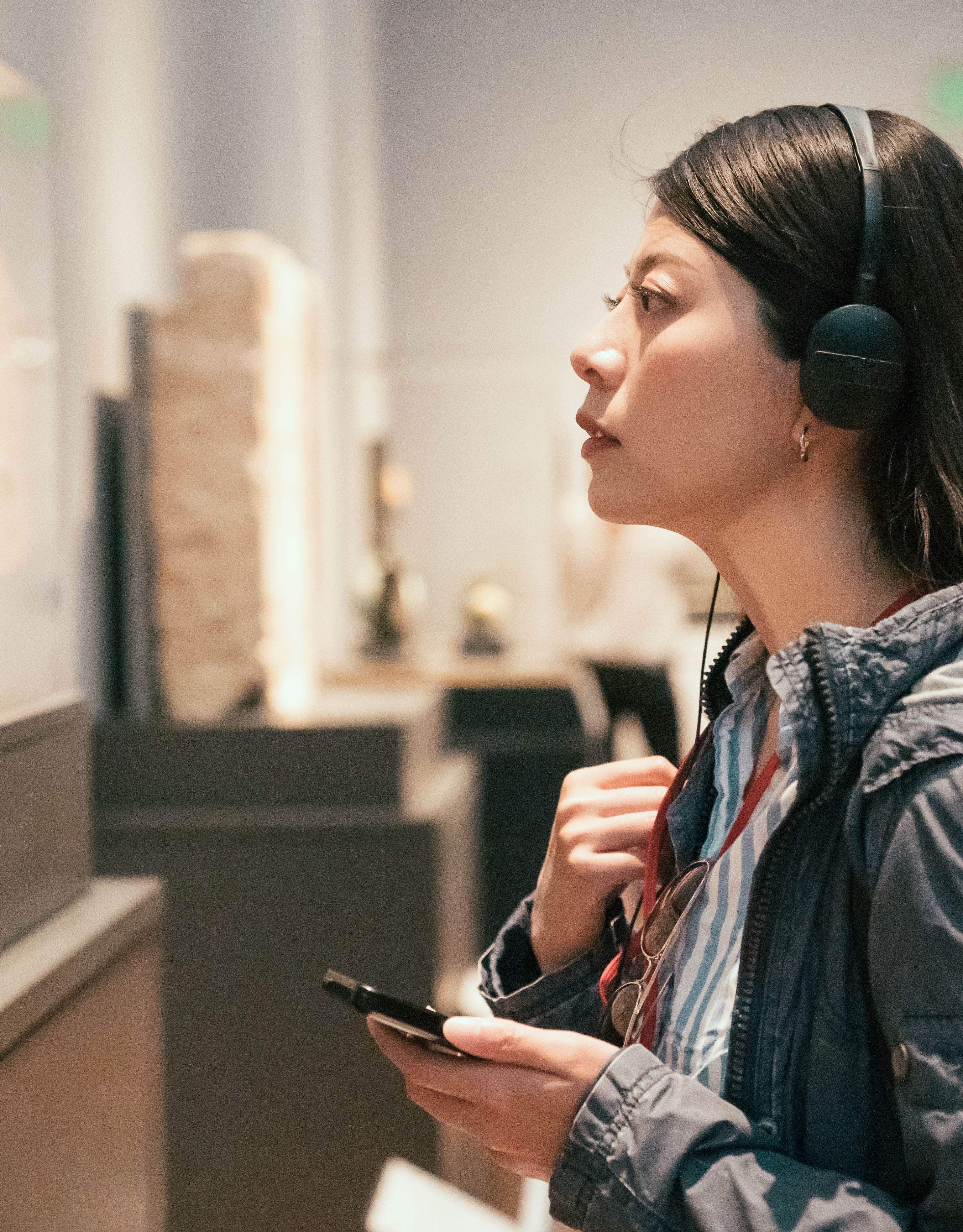 curious young asian chinese woman tourist looking at exposition in museum using headphones and listening audio guide in modern gallery.