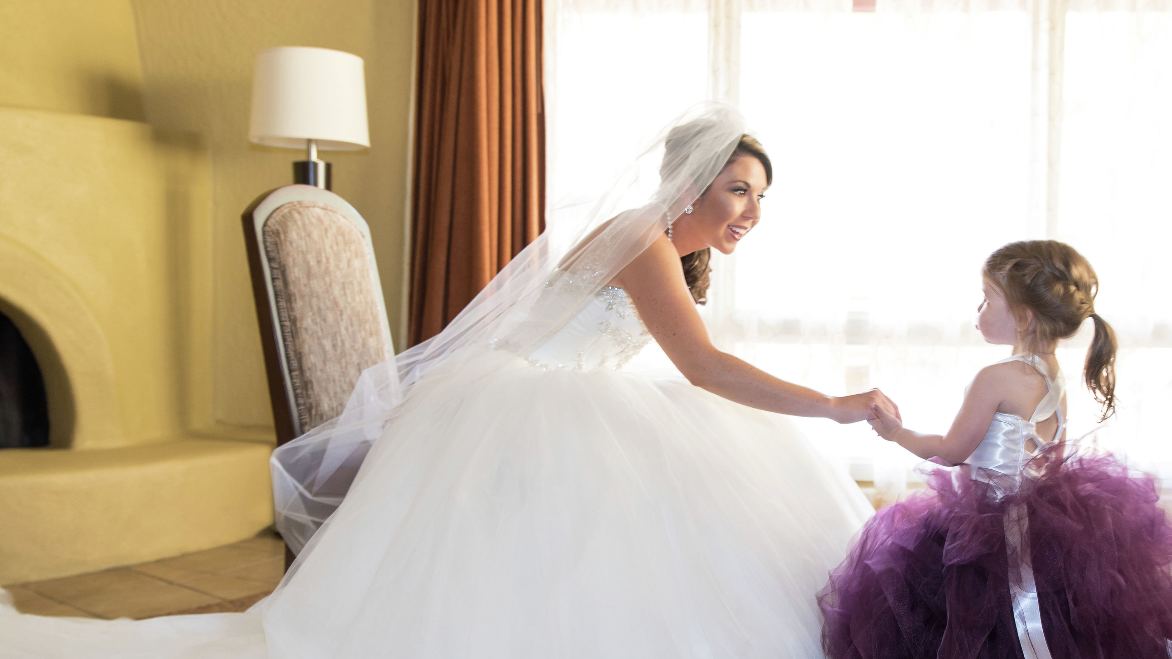 Seated bride in gown holding hand of young flower girl in guest suite