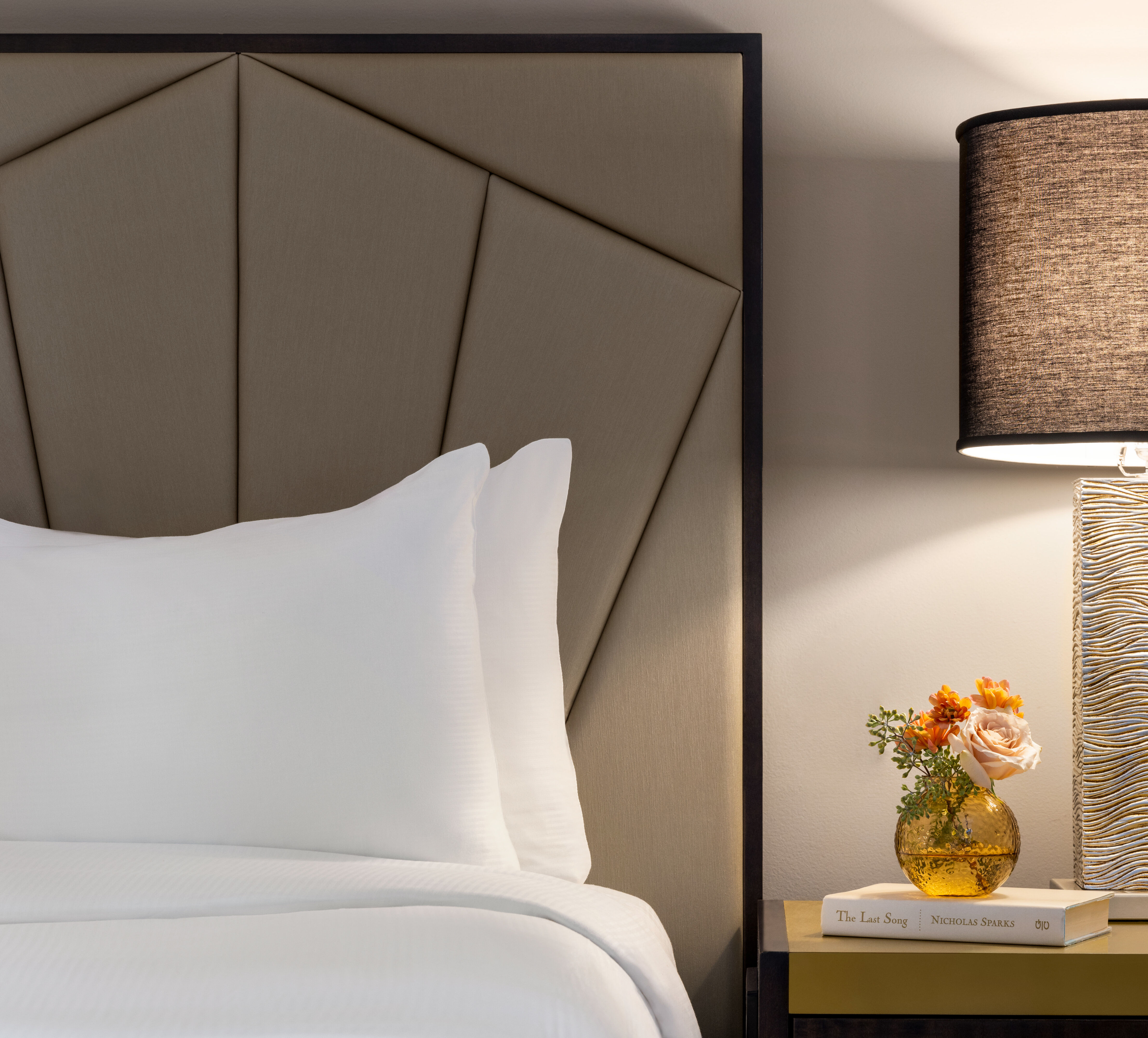 Portion of a bed with white linens, next to a side table with a stone-looking lamp.