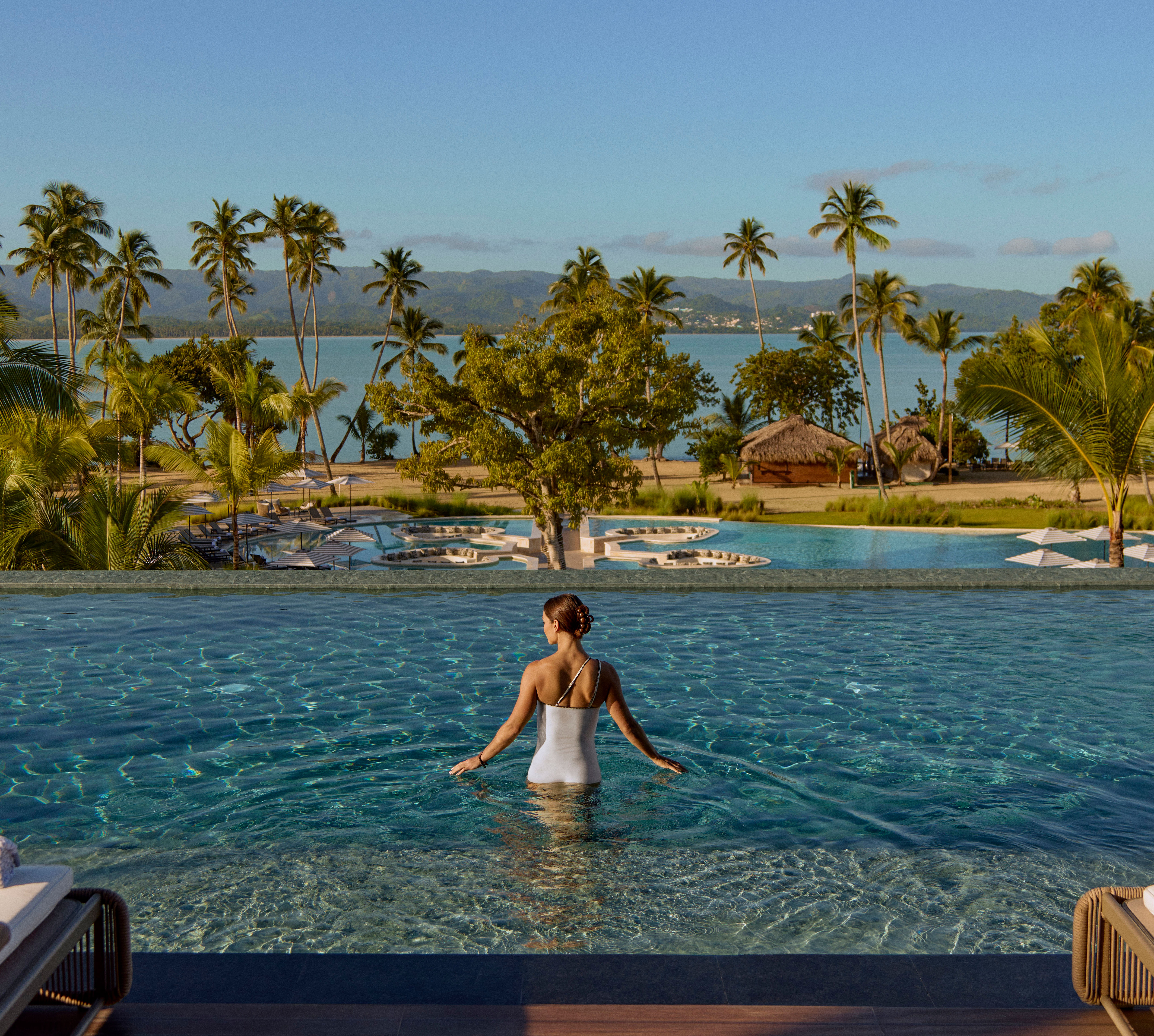 Persoană stând într-o piscină infinită lungă cu vedere la ocean, înconjurată de palmieri, șezlonguri și piscine stratificate într-un peisaj tropical.