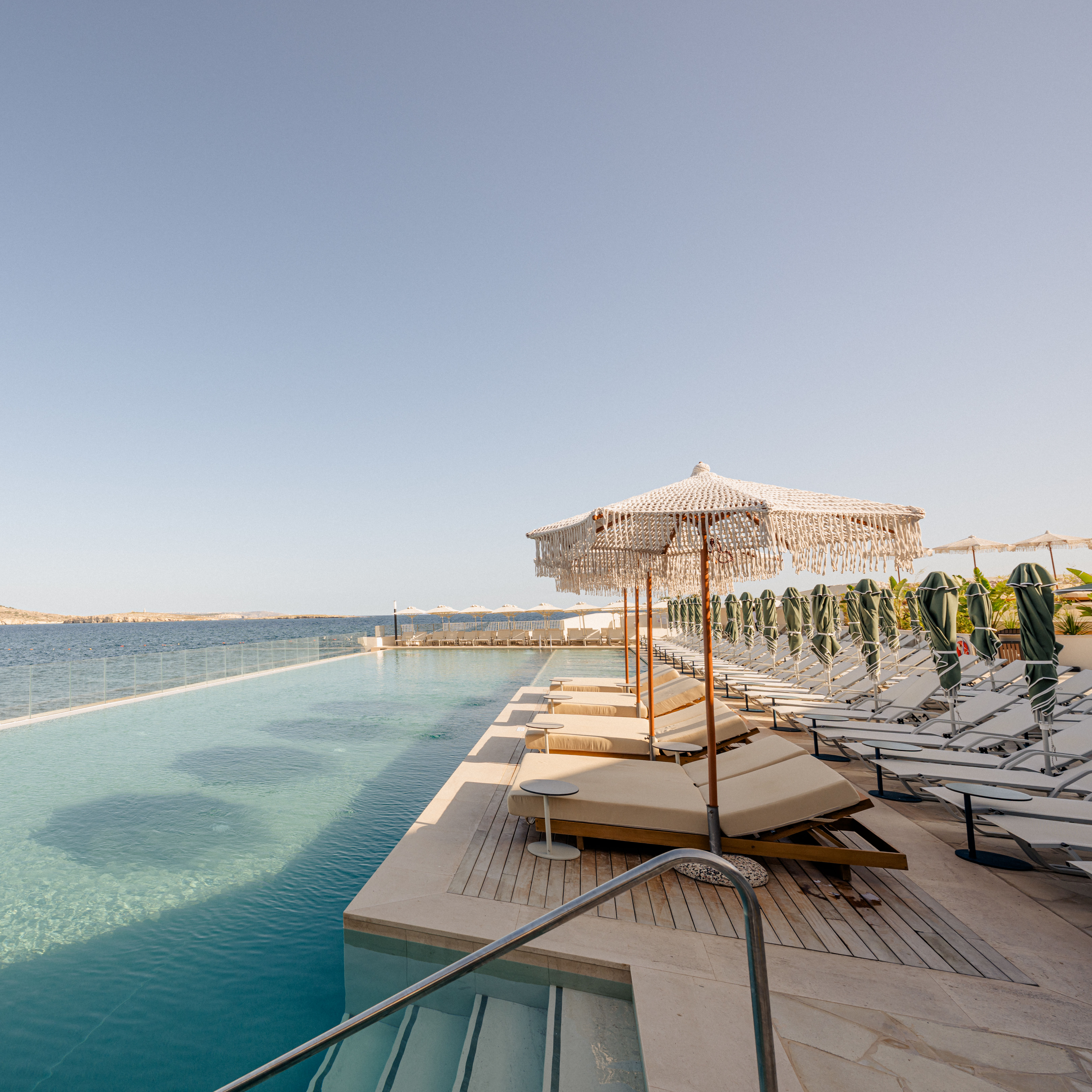 منطقة فناء بها مقاعد للتمدد مطلة على حمام السباحة والمحيط في يوم مشمس