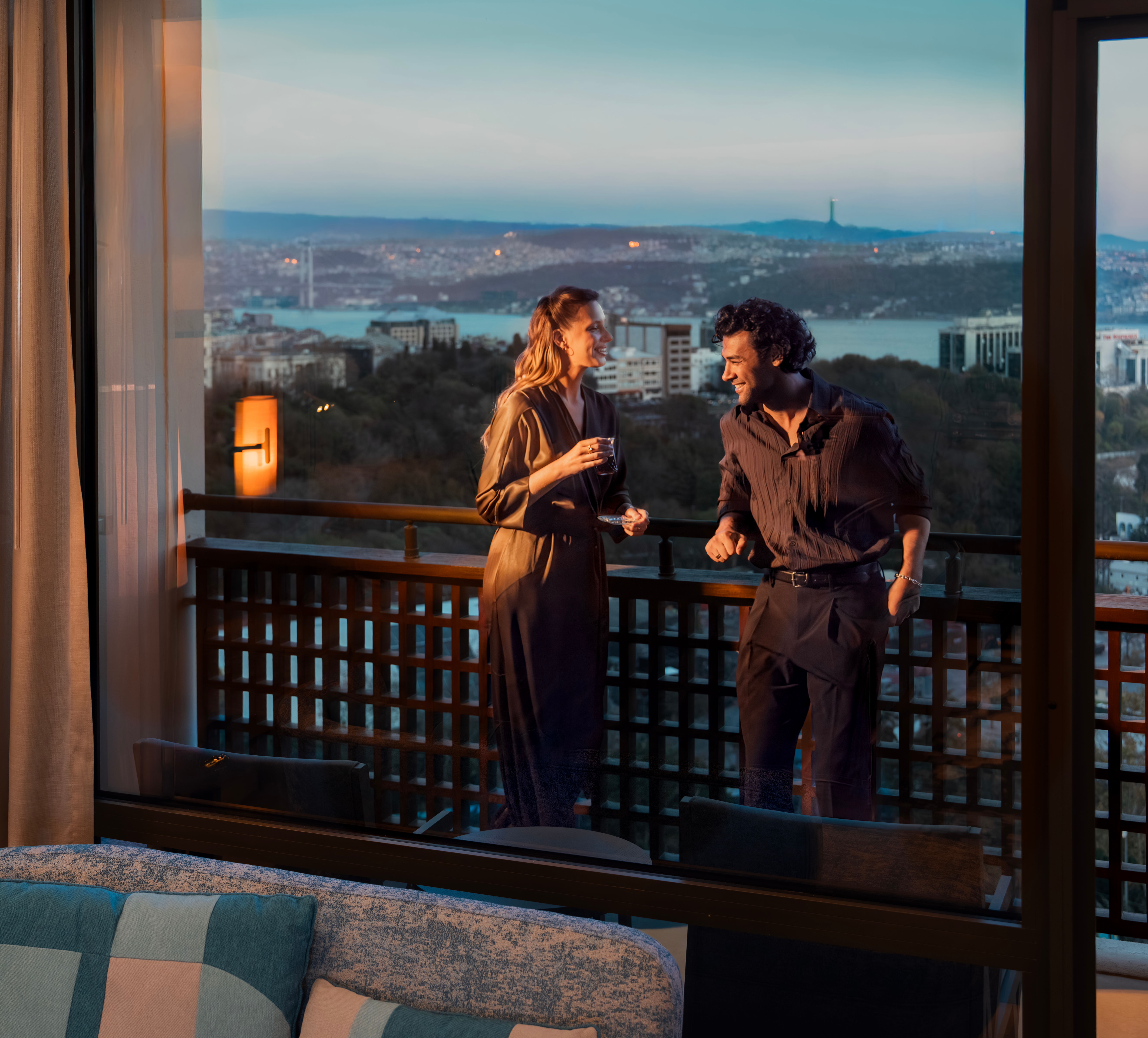 Priviți din interiorul unei camere de hotel printr-o ușă de sticlă către un balcon, unde doi adulți stau cu cerul orașului și apă vizibilă la apus; o canapea, perdele și lampa de podea sunt vizibile în prim-plan.