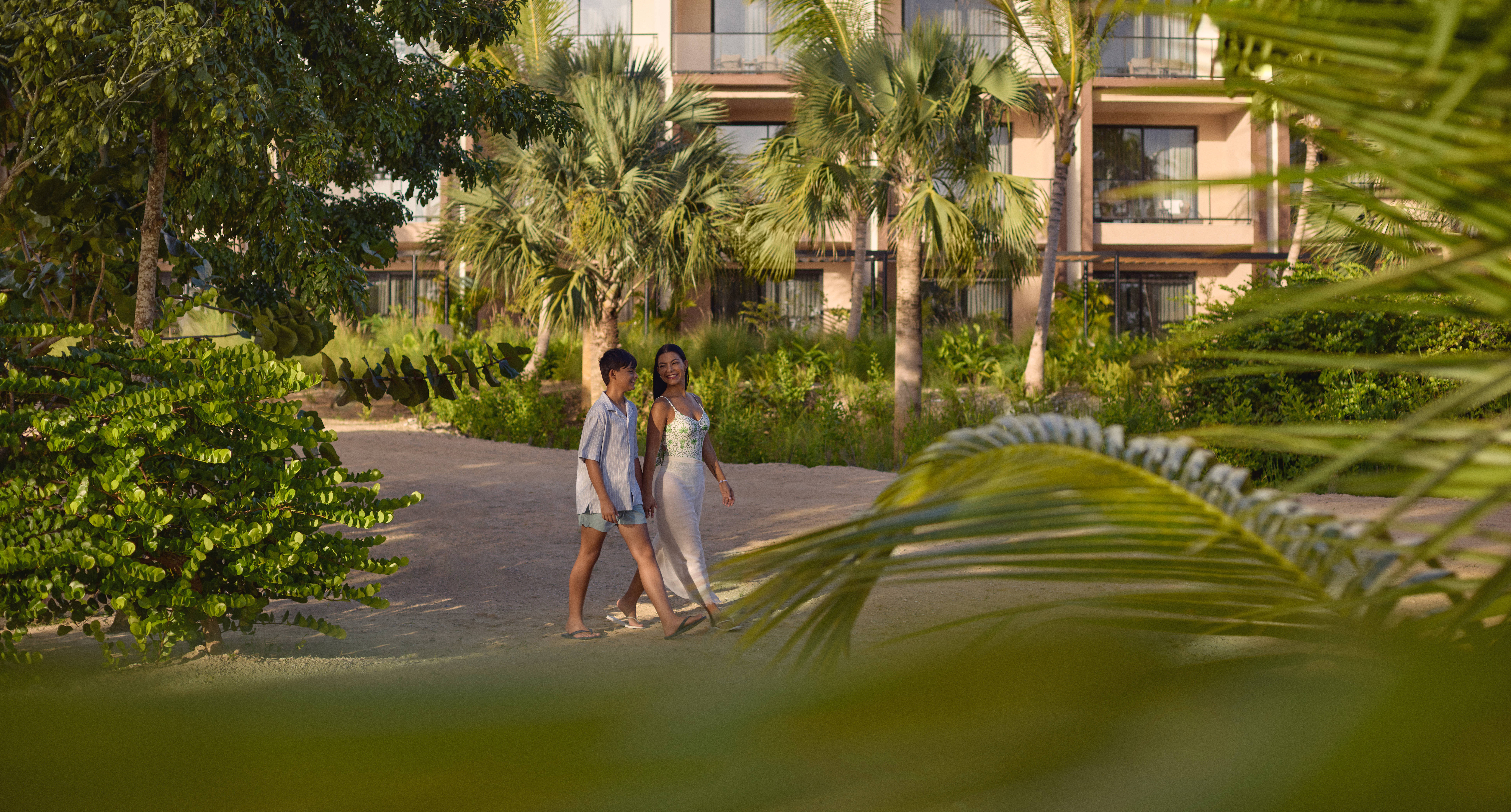Dos personas caminan juntas por un sendero de arena enmarcado por hojas de palmera, con un moderno edificio del resort y exuberante vegetación de fondo.