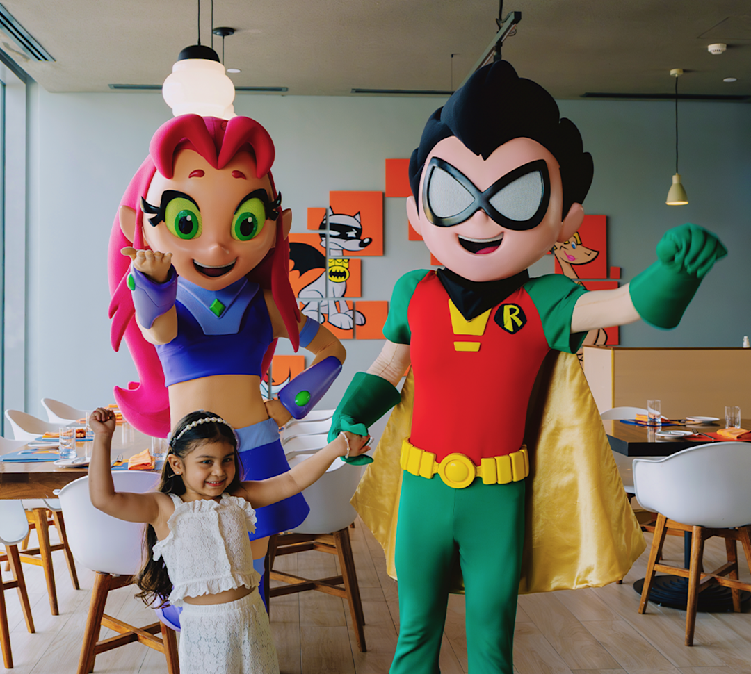 A young girl in a white lace outfit smiles, flexing her arm, between costumed characters of Starfire and Robin from Teen Titans