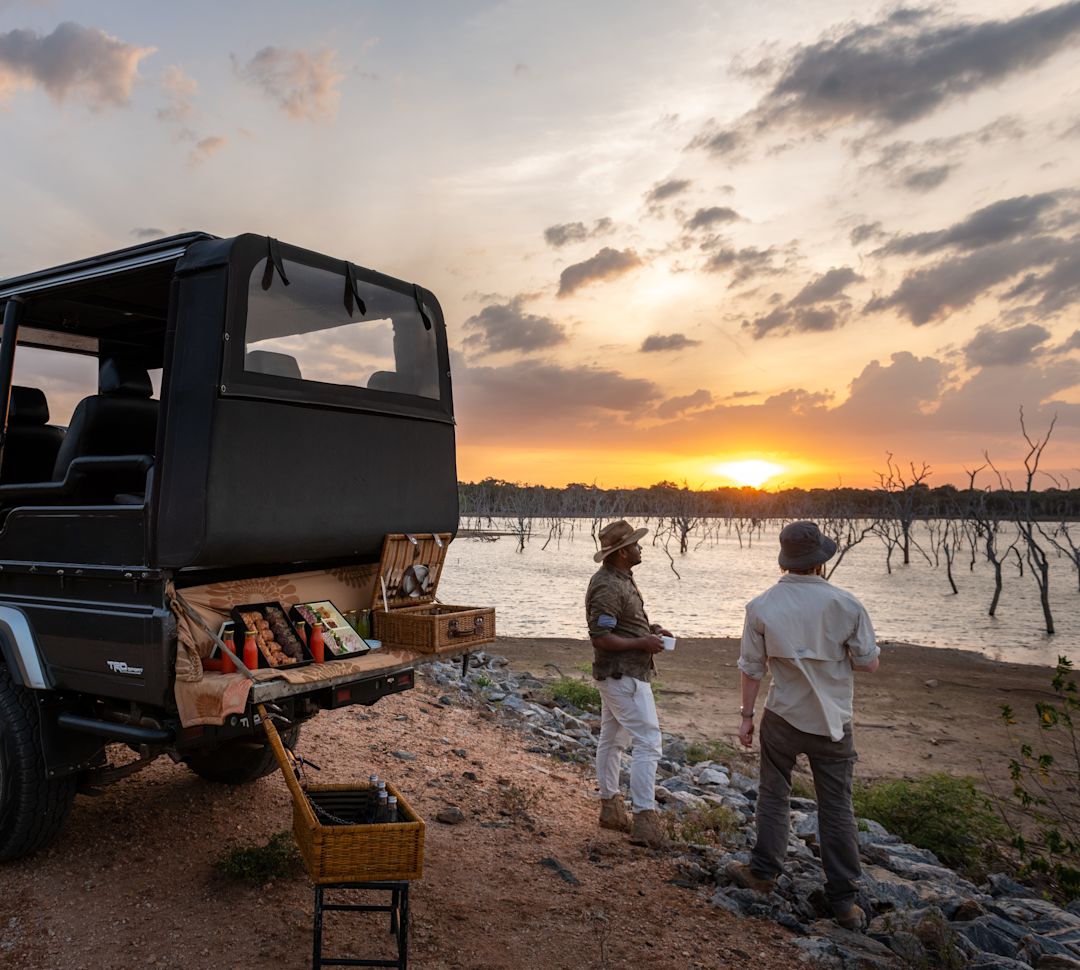 Two men enjoying views of the sunset with picnic on the back of a jeep