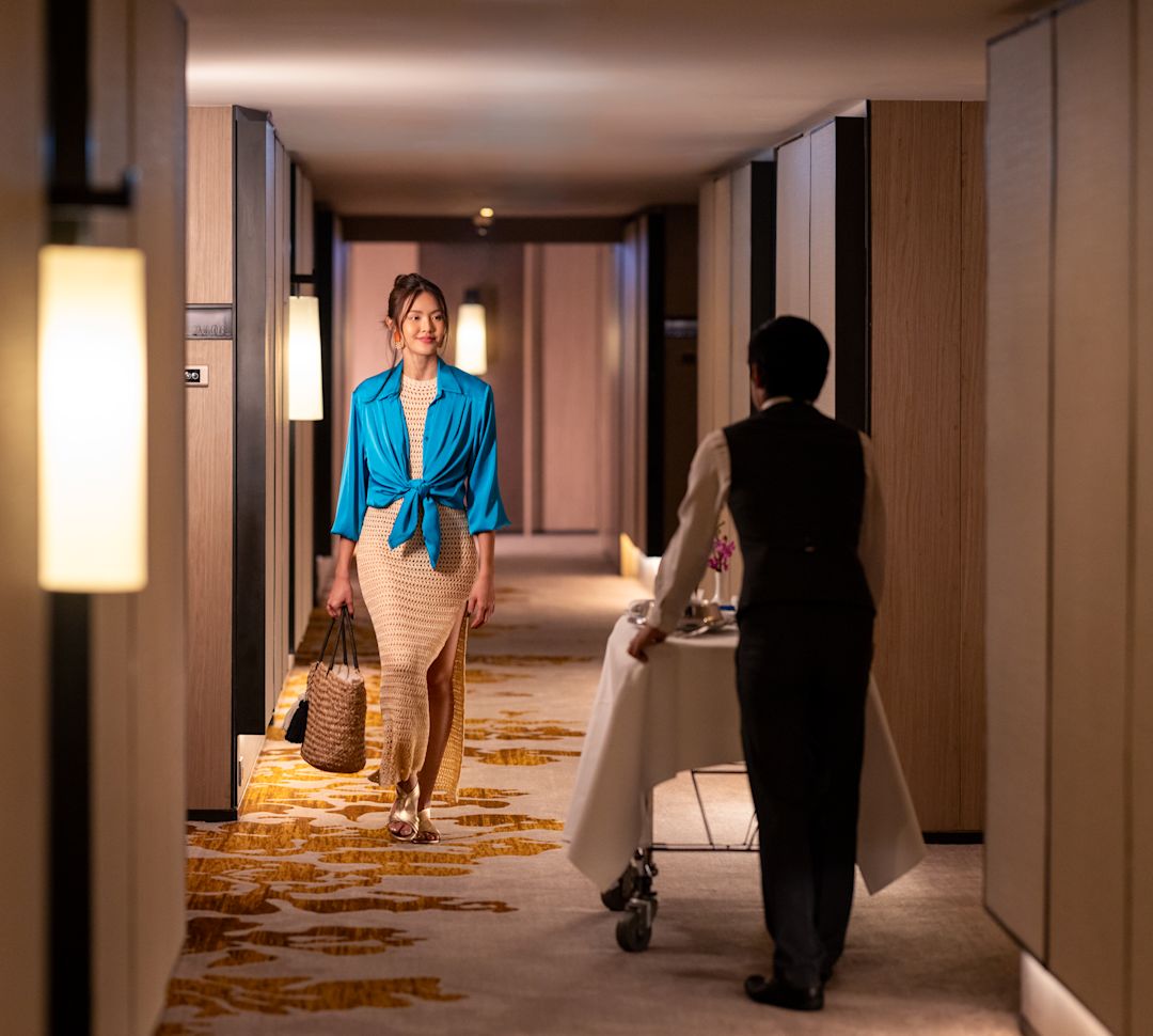 Woman Walking on a Guest Room Hallway while a Staff Member  Delivers Food to a Room