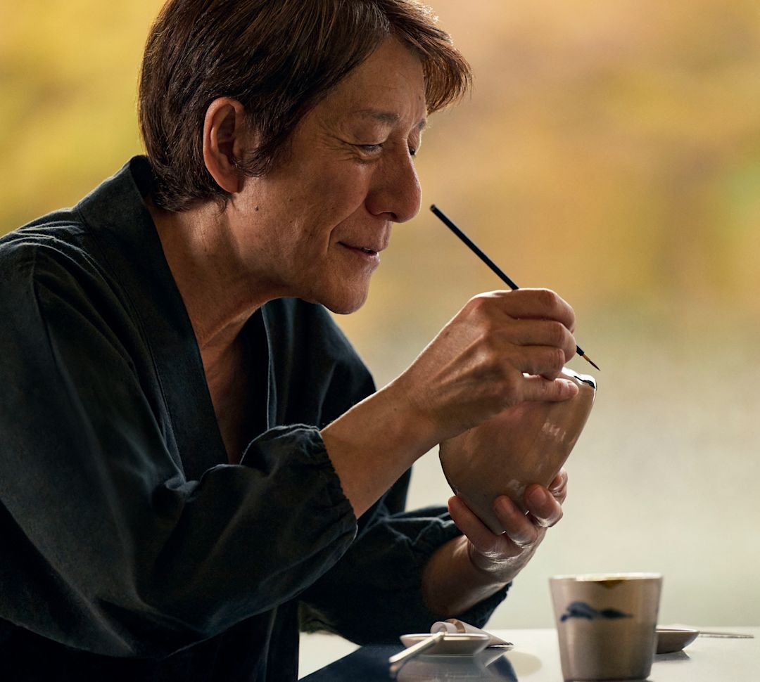 Osoba ćwicząca kintsugi, ostrożnie nakładając lakier na ceramiczną miskę przy stole z małymi narzędziami i filiżankami, z miękkimi jesiennymi kolorami w tle.