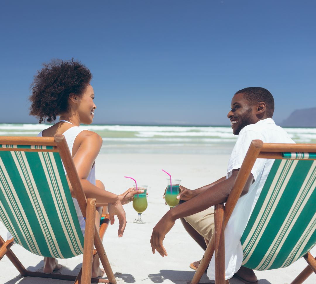 Rear view of happy young multi-ethnic couple relaxing on sun lounger at beach. They are smiling and drinking cocktails.