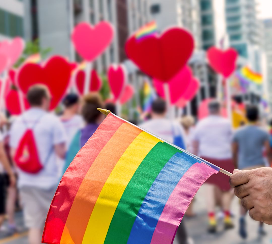 Hand holds a pride flag in front of a street of people with heart shaped signs