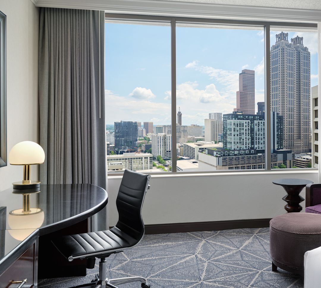 King room city view with edge of bed and view of window with table and purple chair-transition