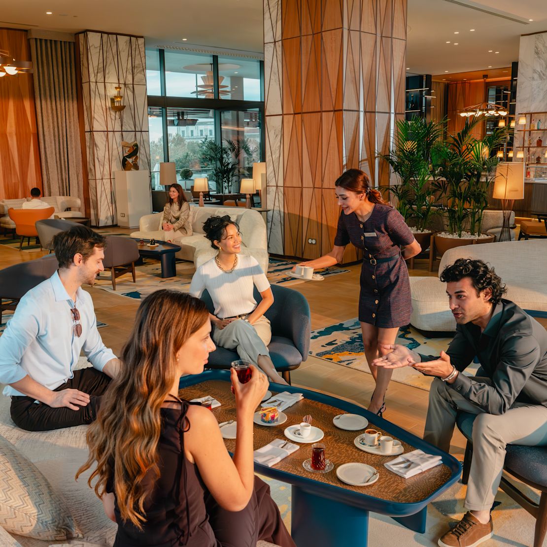 Un grup de adulți așezați într-un lounge spațios din holul hotelului, în jurul meselor joase, cu ceai și deserturi, în timp ce un membru al personalului pune obiecte pe masă; scaunele tapițate, plantele și lumina caldă umplu spațiul.- Tranziția