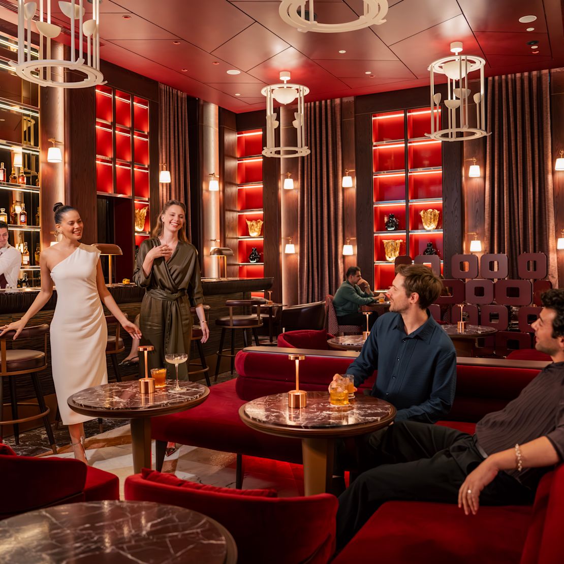 Vedere interioară a unui bar sau a unui lounge dintr-un hotel cu scaune roșii tapițate, mese din marmură, adăpost iluminat în spatele barului și mai mulți adulți așezați și stând în picioare în spațiu.- Tranziție