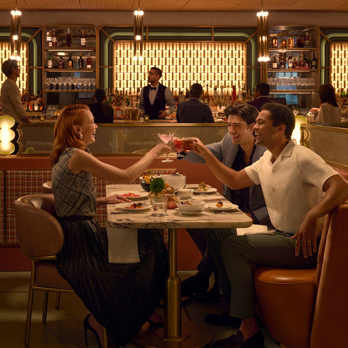 Une femme et deux amis hommes savourent des boissons et des collations légères au Lex Yard. En arrière-plan, d'autres clients et un appel d'offres au bar peuvent être vus en train de parler à un client au bar. -transition