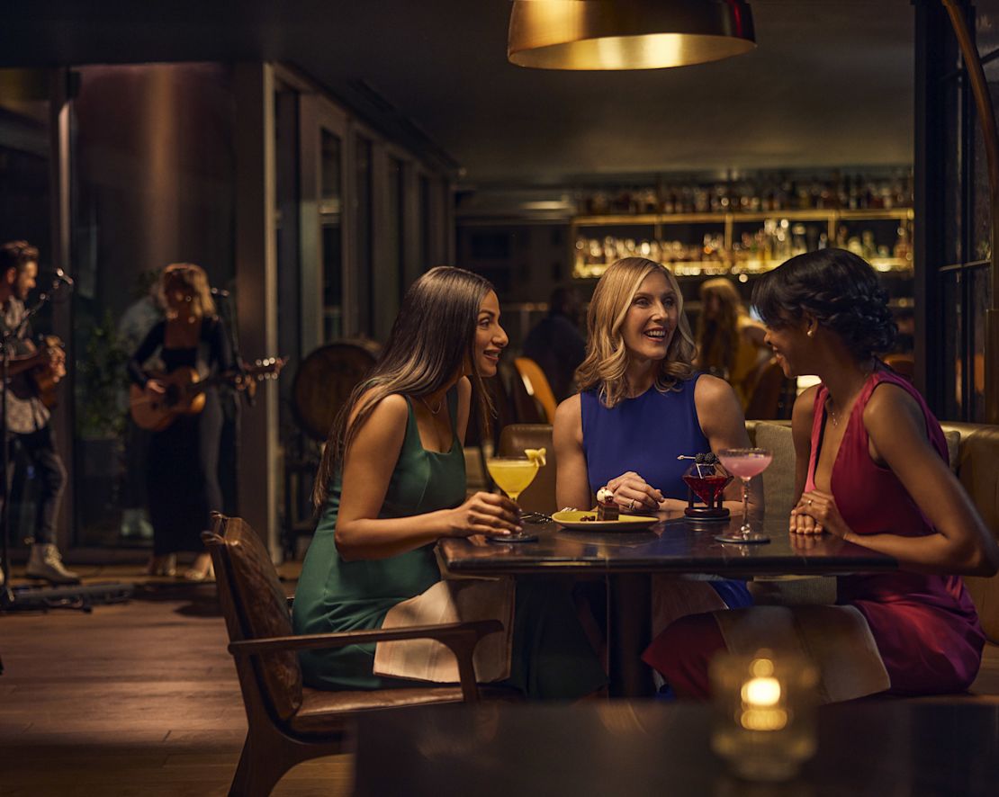 Three women smiling in seating area while live music plays behind-transition