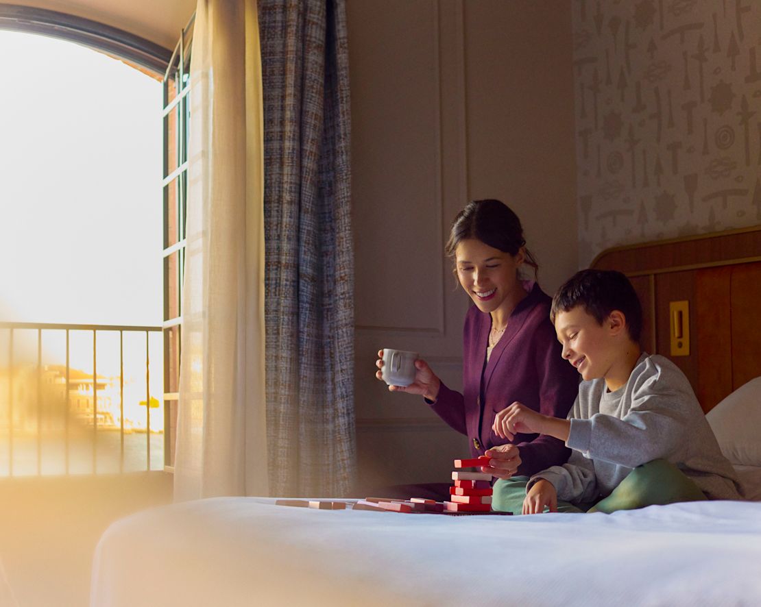 Son playing with a wooden puzzle on the bed while mother watches and enjoys a cup of coffee or tea-transition
