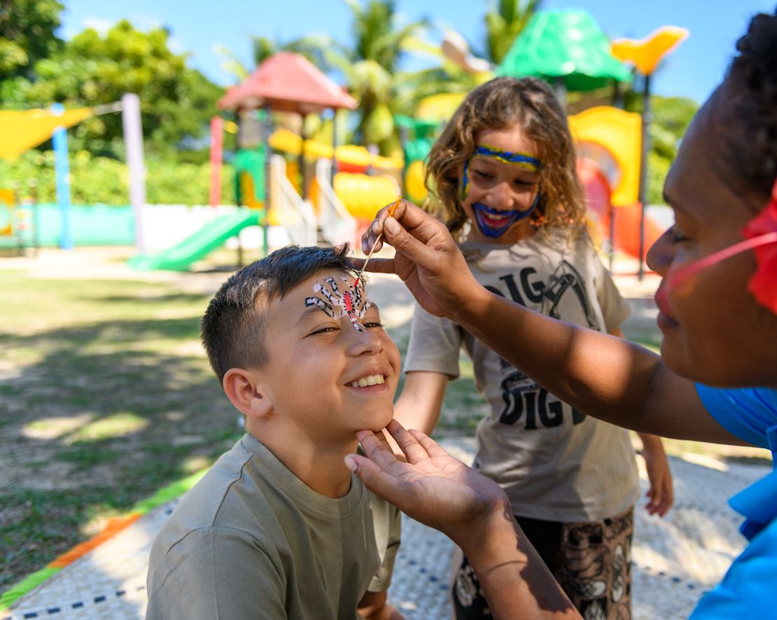 Člen týmu maluje tvář mladého chlapce s malou dívkou, která se dívá na dětský klub.-přechod