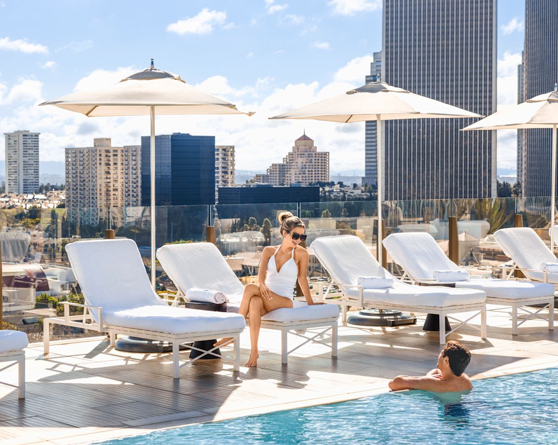 女子坐在躺椅上，男子在泳池中放松，背景为城市景观过渡