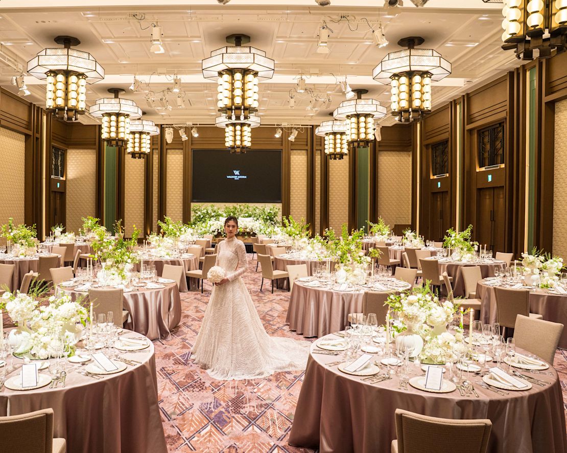 Mireasă stând într-o sală de bal decorată cu flori albe în mese rotunde pentru a sărbători o tranziție de nuntă