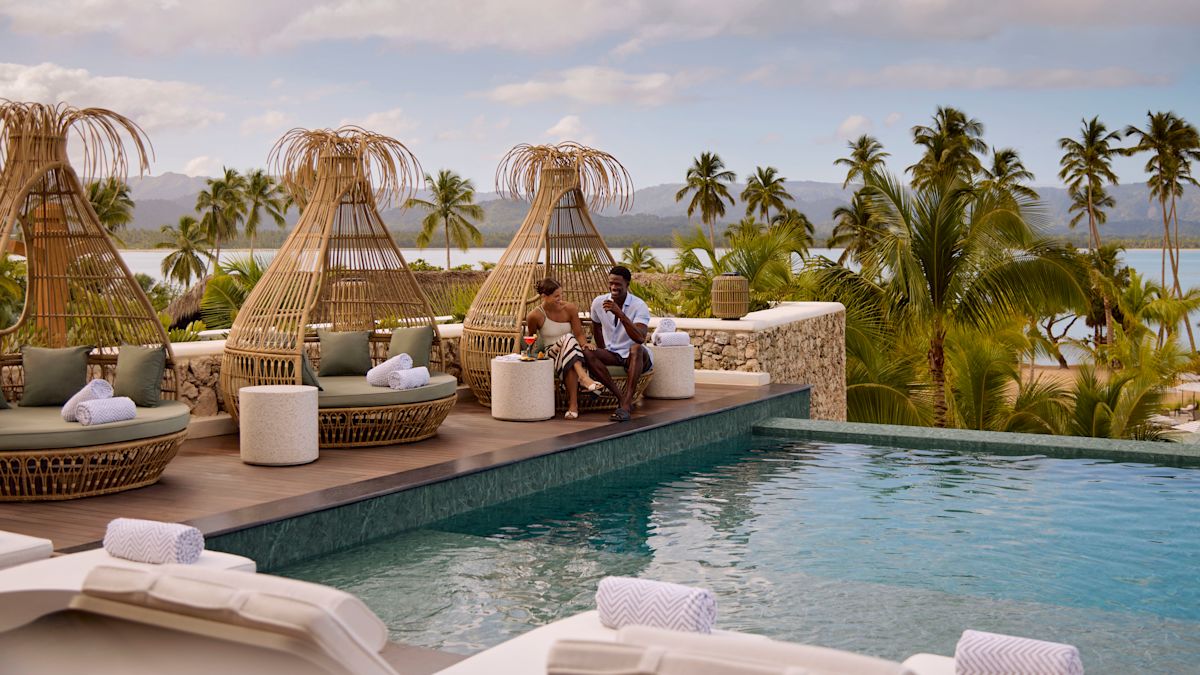 Doi oameni relaxându-se lângă o piscină ridicată, pe păstăi de lounge țesute, înconjurate de palmieri și priveliști asupra oceanului și dealurilor îndepărtate, sub cerul parțial înnorat.