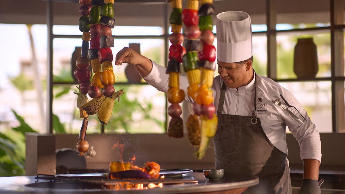 Chef tending to skewers of colorful vegetables over an open flame at an outdoor grill station.
