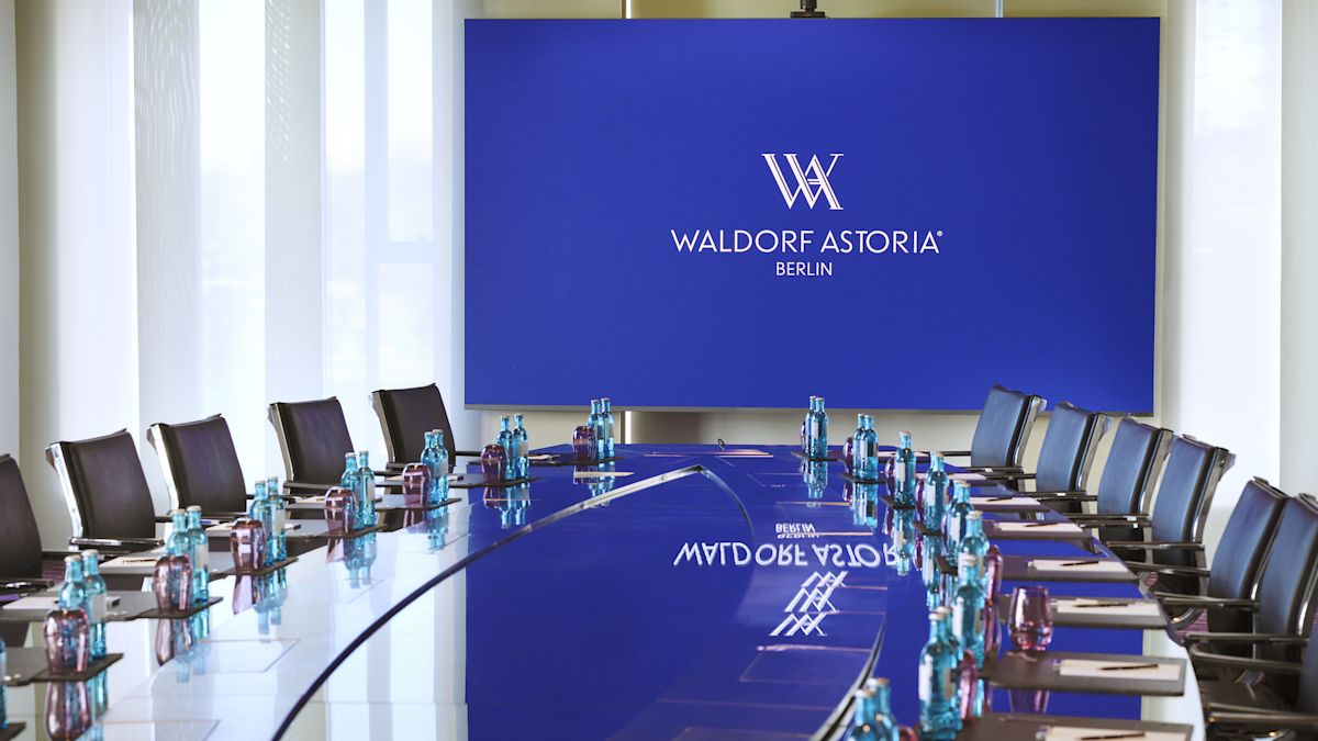 stůl konferenční místnosti s jídelním prostorem, láhvemi na vodu, modrou obrazovkou s textem a logem Waldorf Astoria Berlin