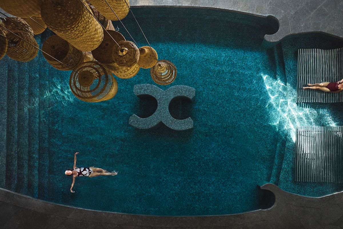 Pemandangan mata burung dari seorang wanita yang mengambang di punggungnya di kolam renang sambil bersantai sementara wanita lain beristirahat di salah satu lounger di kolam renang