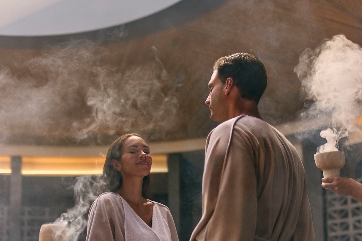 Seorang wanita dan seorang pria bergandengan tangan saat berdiri di spa dan anggota tim memegang mangkuk dengan dupa rokok