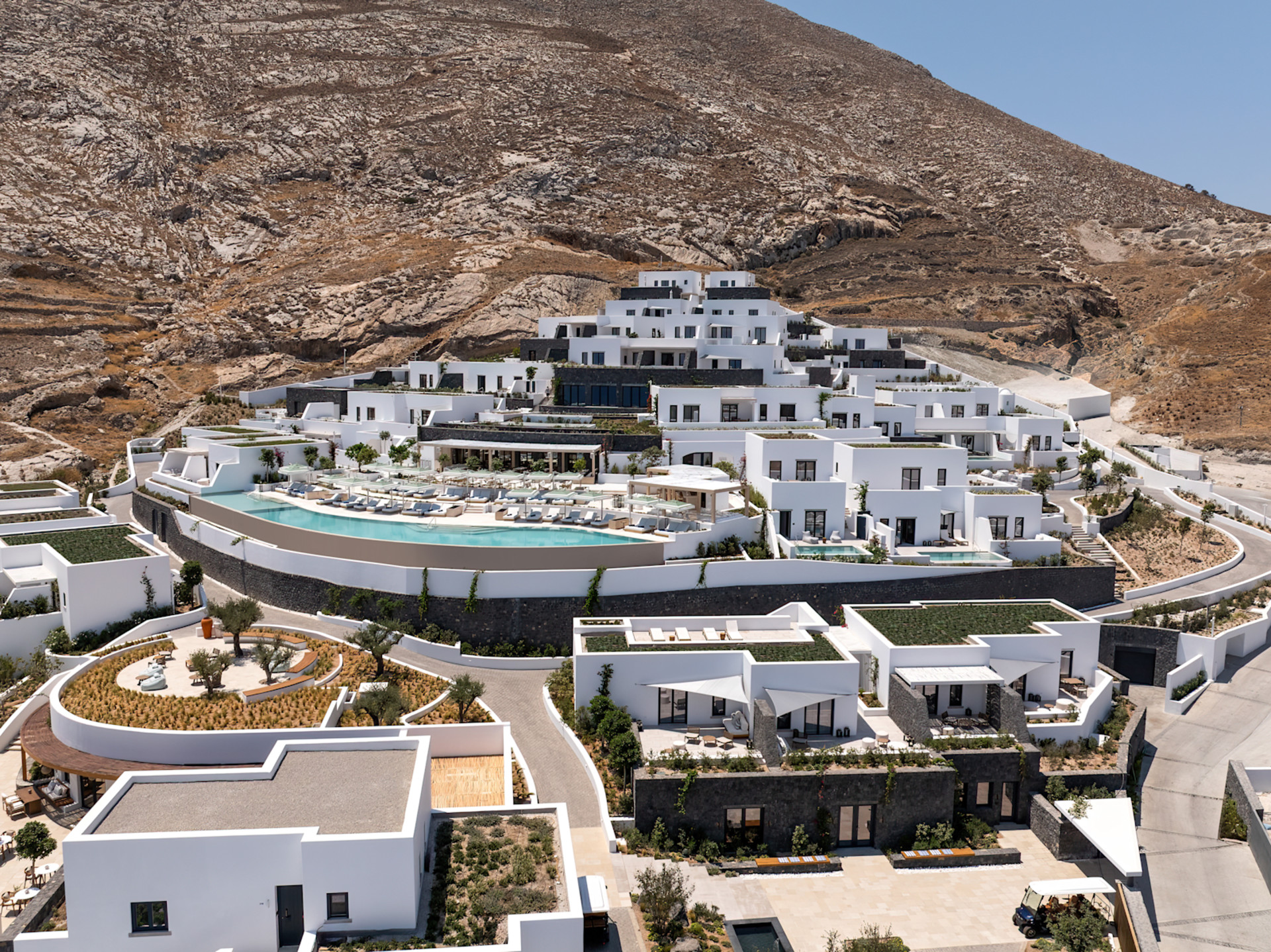 Gün boyunca beyaz boyalı duvarları ve çok sayıda açık oturma alanı bulunan açık havuzu gösteren binanın dış görünümü.