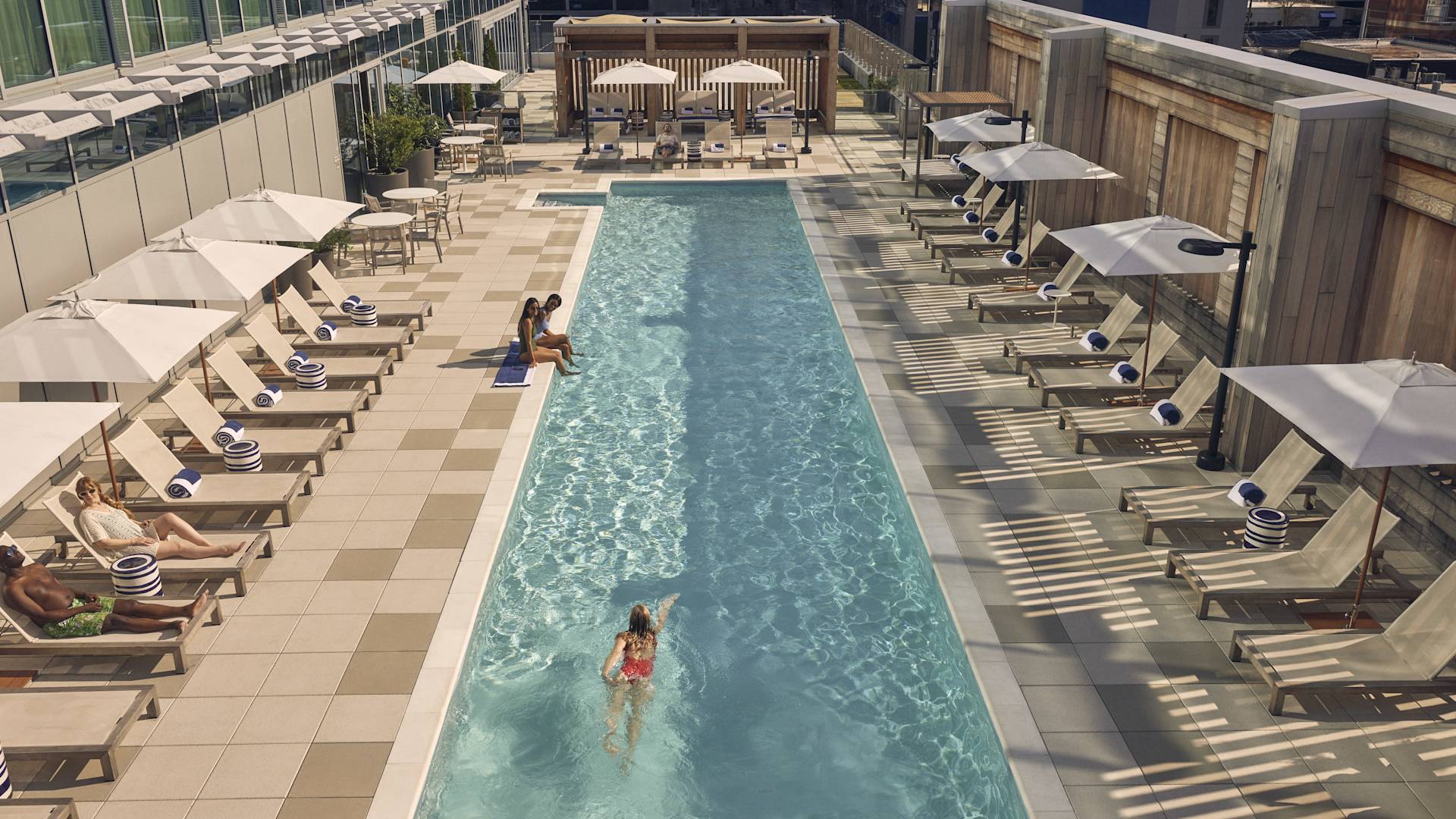 Two woman relax at the side of the pool, while woman in red swimsuit does lengths