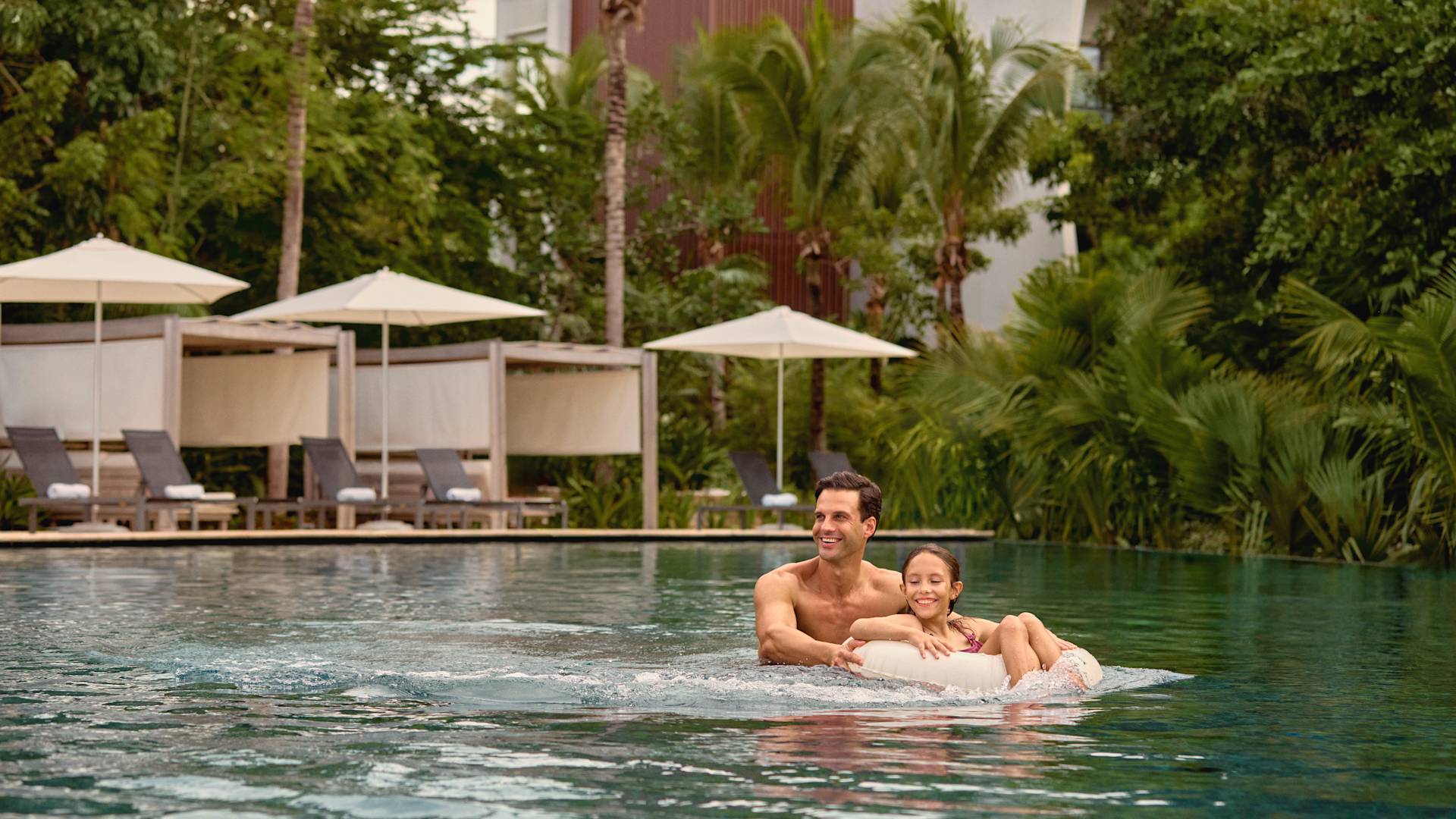 아버지가 수영장의 딸을 공기주입식 도넛으로 밀고 있습니다.