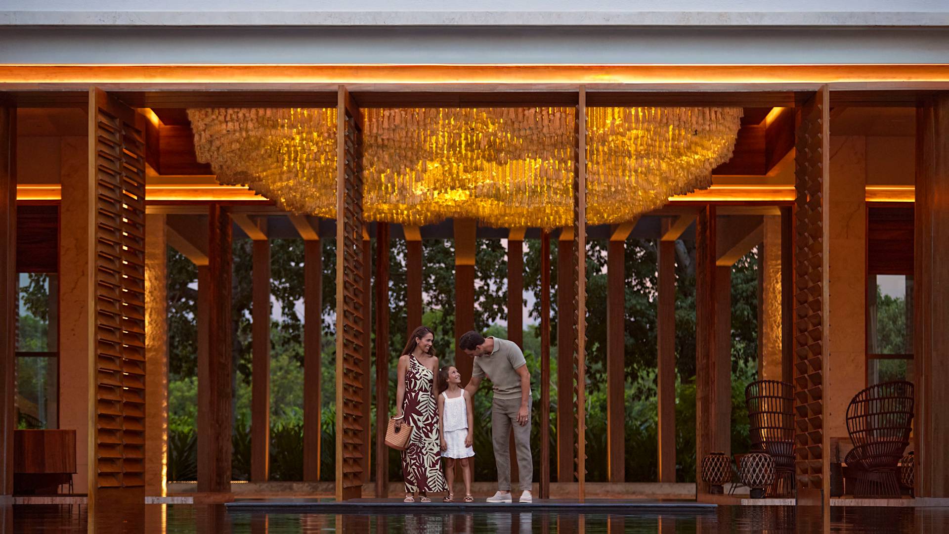 Äiti, isä ja tytär seisovat suuren vesiominaisuuden vieressä valaistussa ja osittain ulkoilmahotellin aulassa