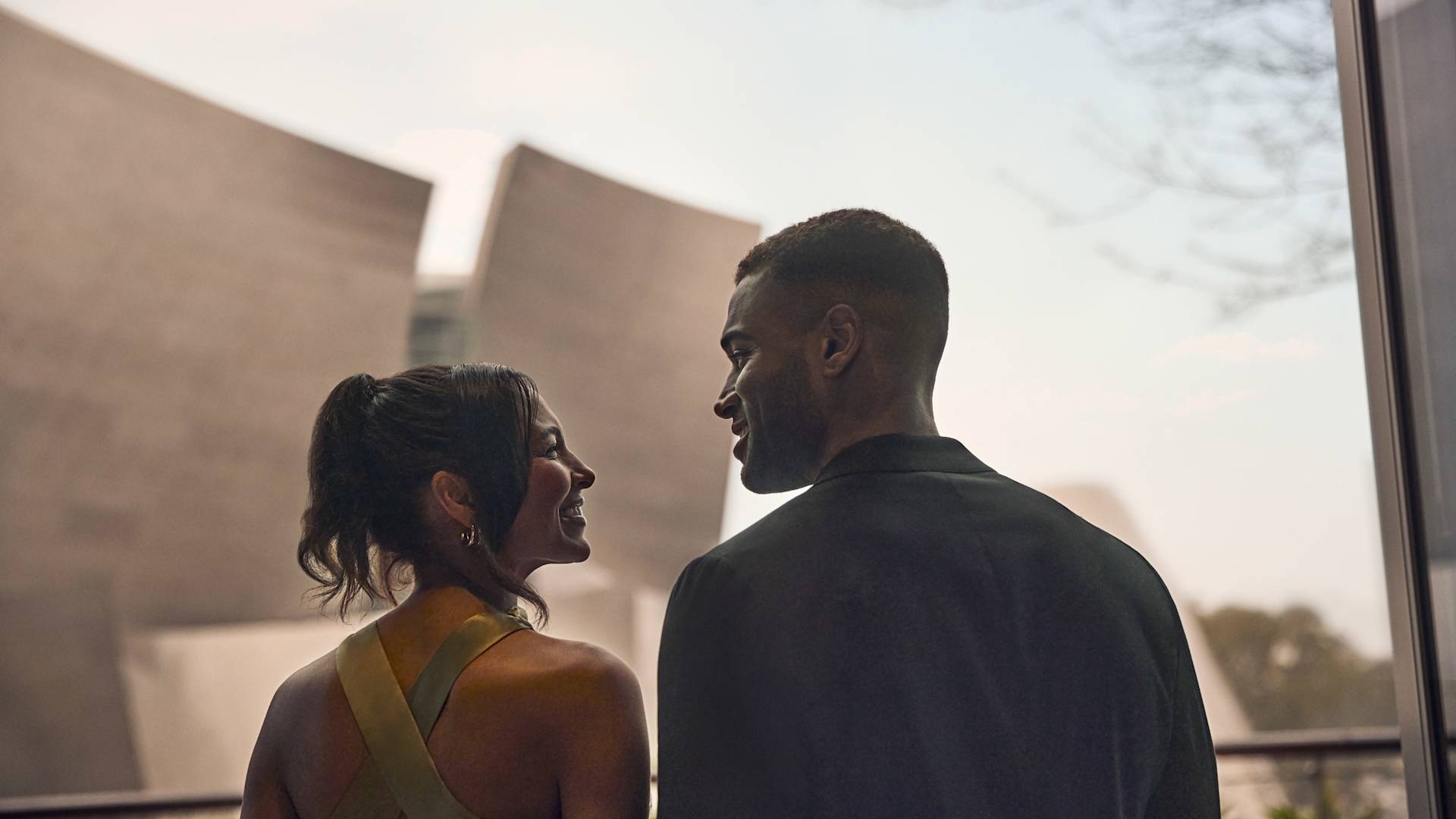 Homem e mulher sorrindo um para o outro, com o exterior do hotel ao fundo