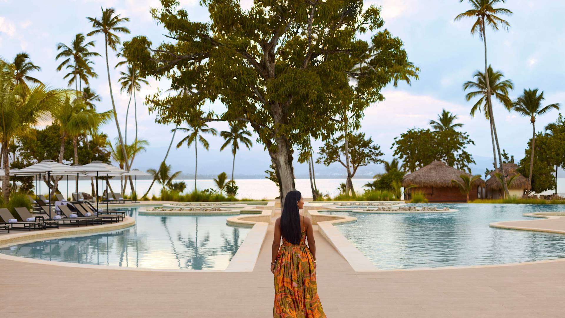 Persoană care merge către un copac mare în centrul unei zone liniștite din piscină, înconjurată de palmieri, scaune de relaxare și priveliști către ocean la apus.