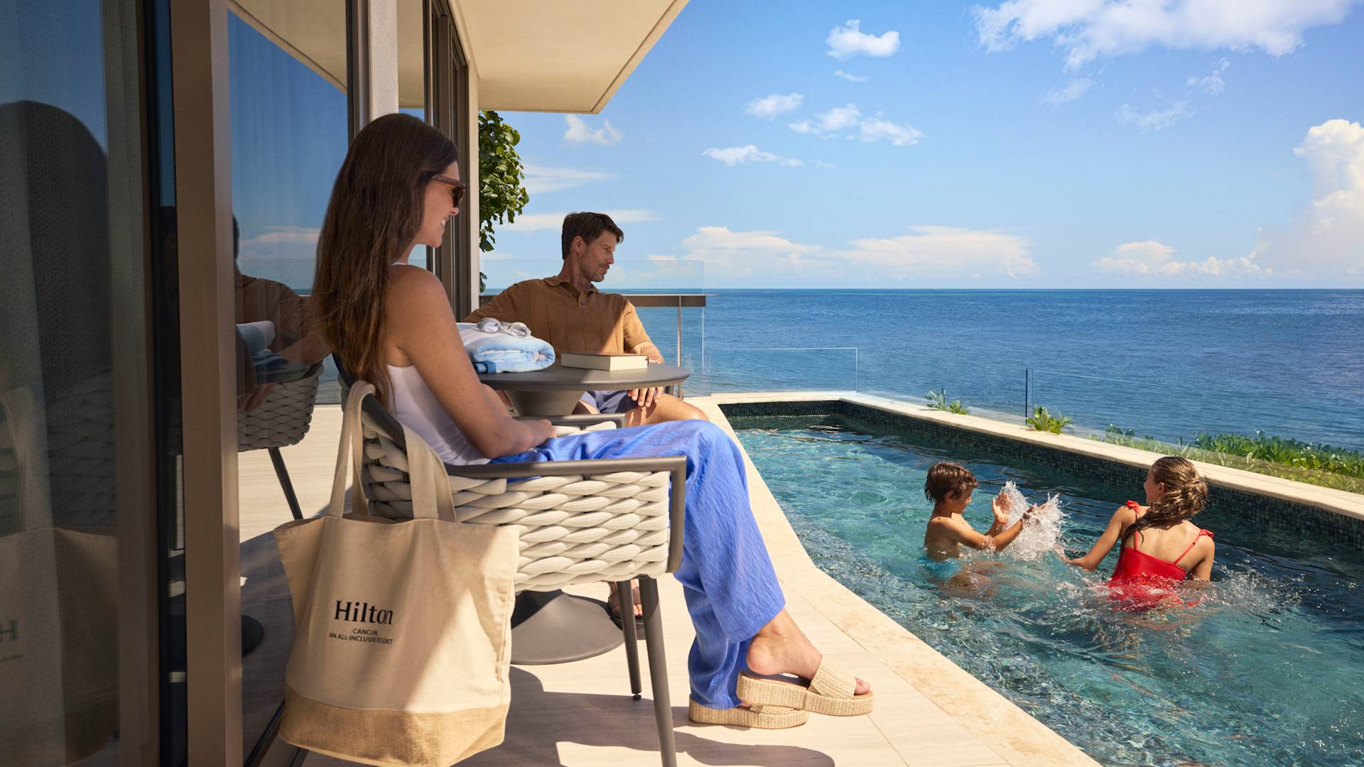 Mamă și tată relaxându-se pe terasa cu vedere spre mare în timp ce fiul și fiica lor stropesc în piscină