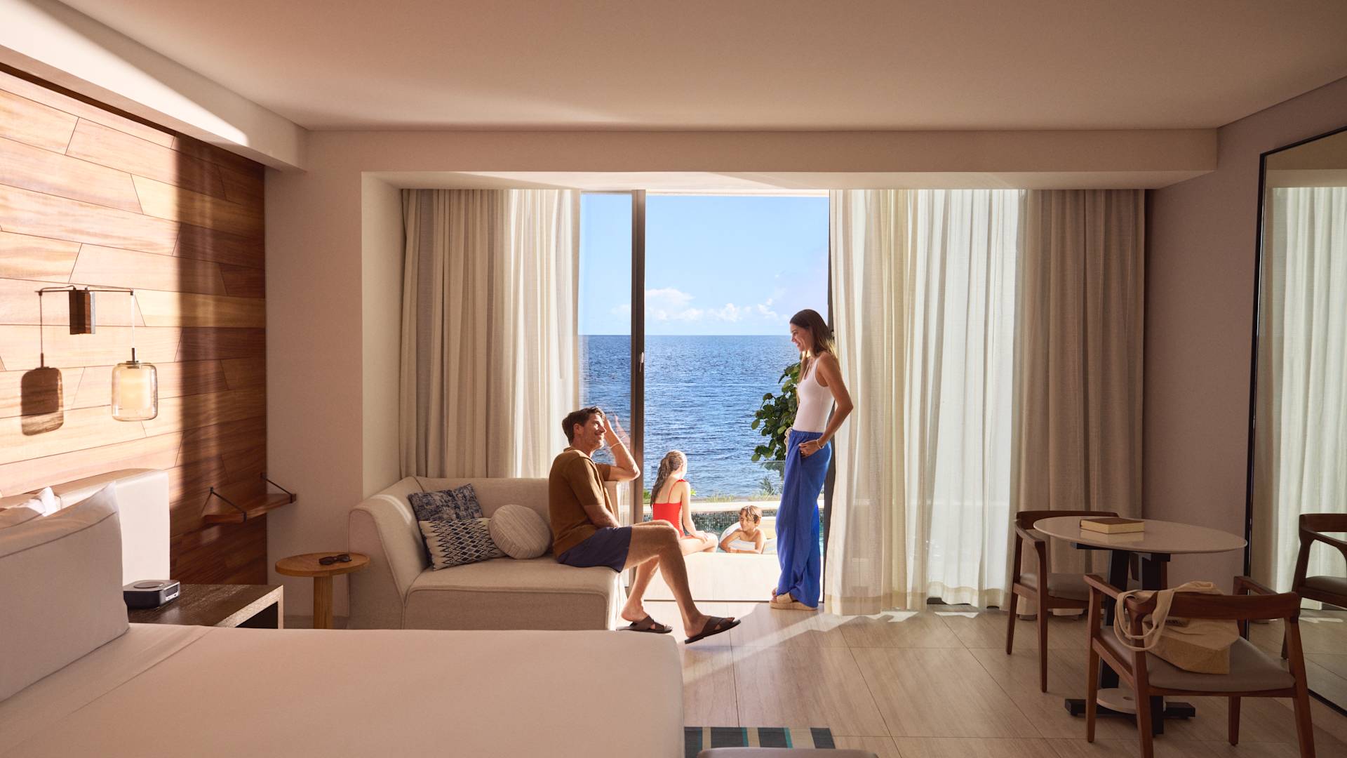 Mother and father relaxing and talking in a modern beachfront hotel room while the daughter and son are relaxing outside