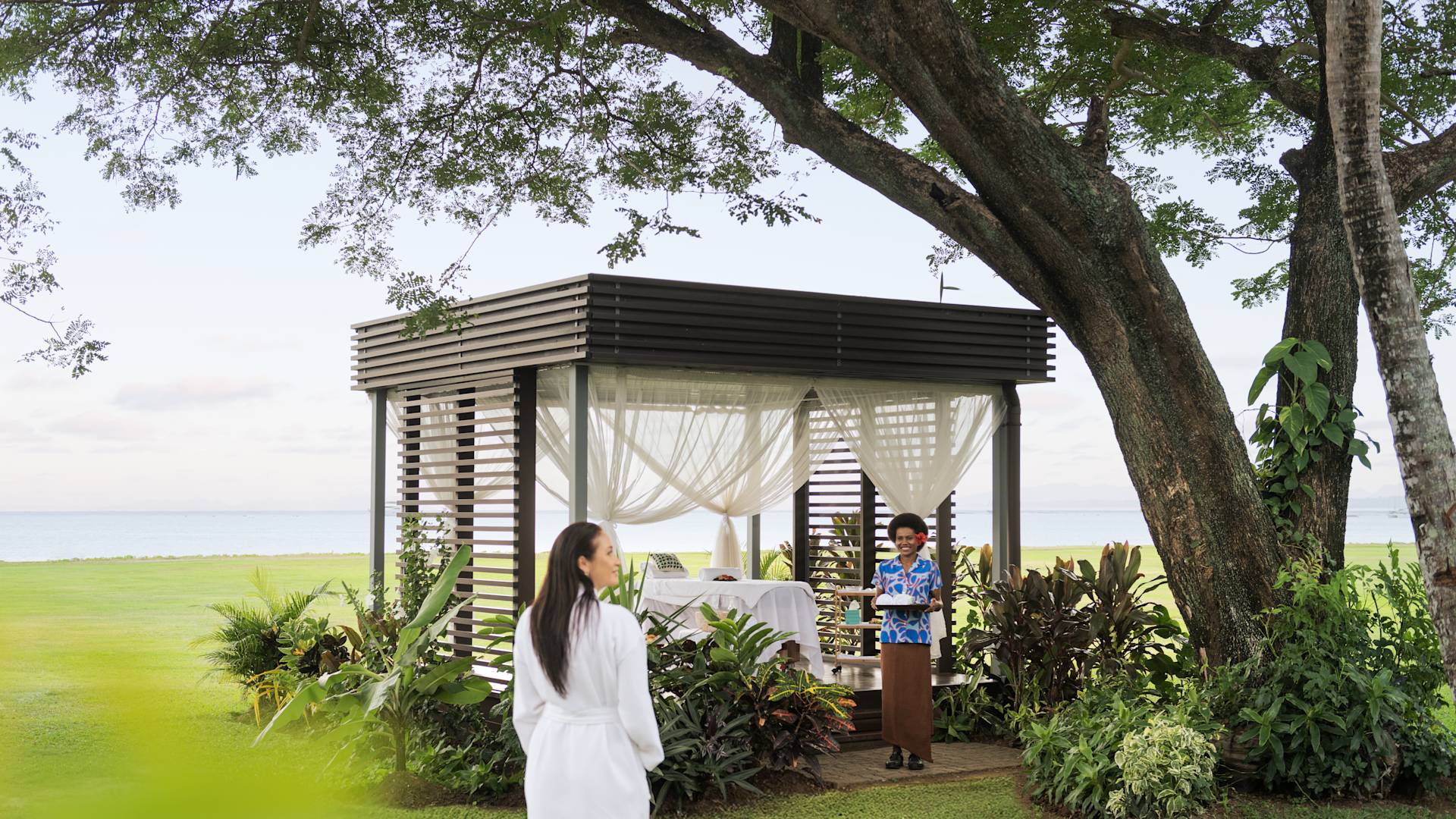 Woman approaching a spa treatment set up under a tree, and a female team member waiting to receive her.