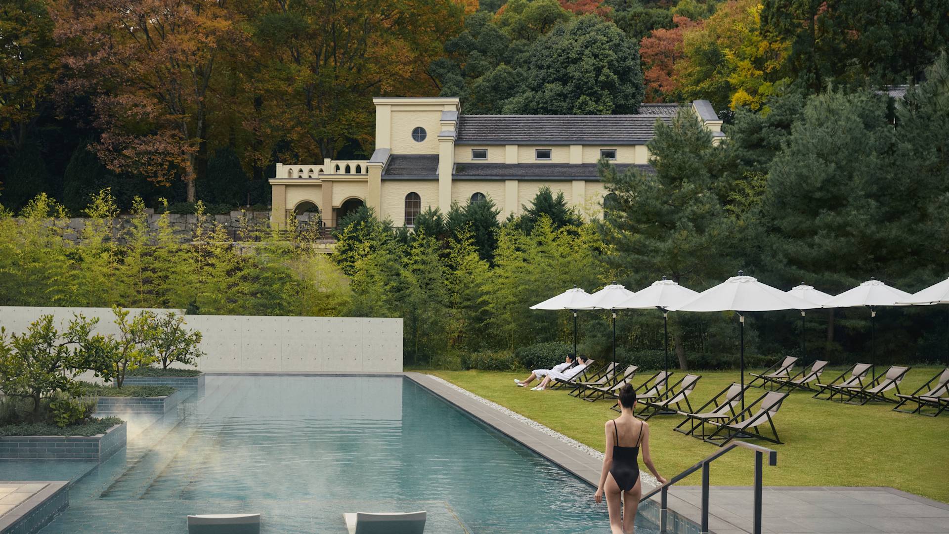Persona che entra in una piscina esterna con vapore circondata da sedie a sdraio, ombrelloni e lussureggianti alberi autunnali, con un edificio in stile storico sullo sfondo.