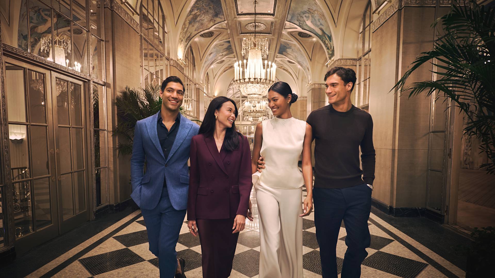 Four elegantly dressed people, two men and two women, walk together down a grand hotel corridor with a checkered floor, ornate chandeliers, and a high, arched ceiling with murals.