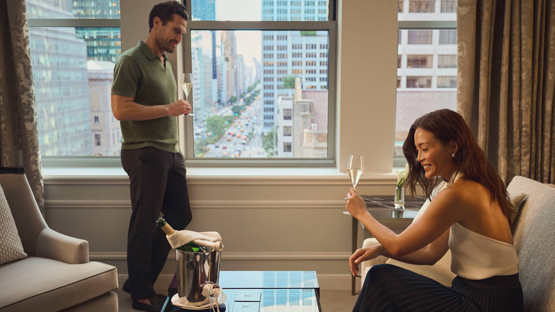 Man and woman enjoying a bottle of champagne in their hotel room. Both are wearing smart-casual clothes and are sharing a nice  relaxed moment with each other. Through the window, the streets of New York are visible.