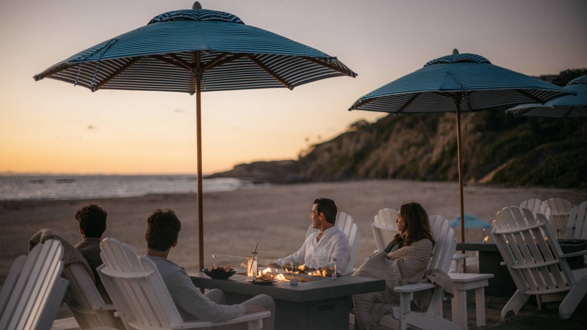 a group of people sitting around a fire pit and having drinks at the beach