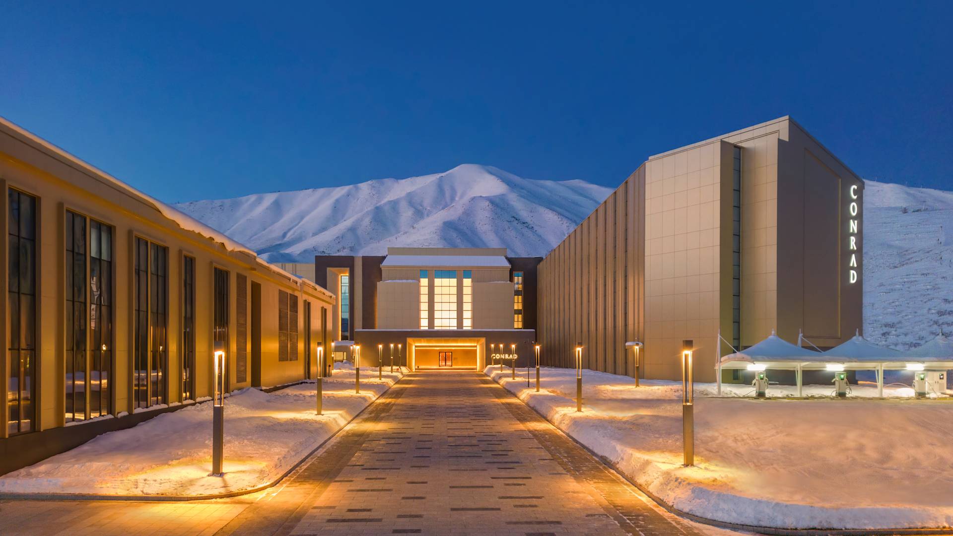 Vstup do hotelu s dlouhou příjezdovou vozovkou, osvětlením a horami