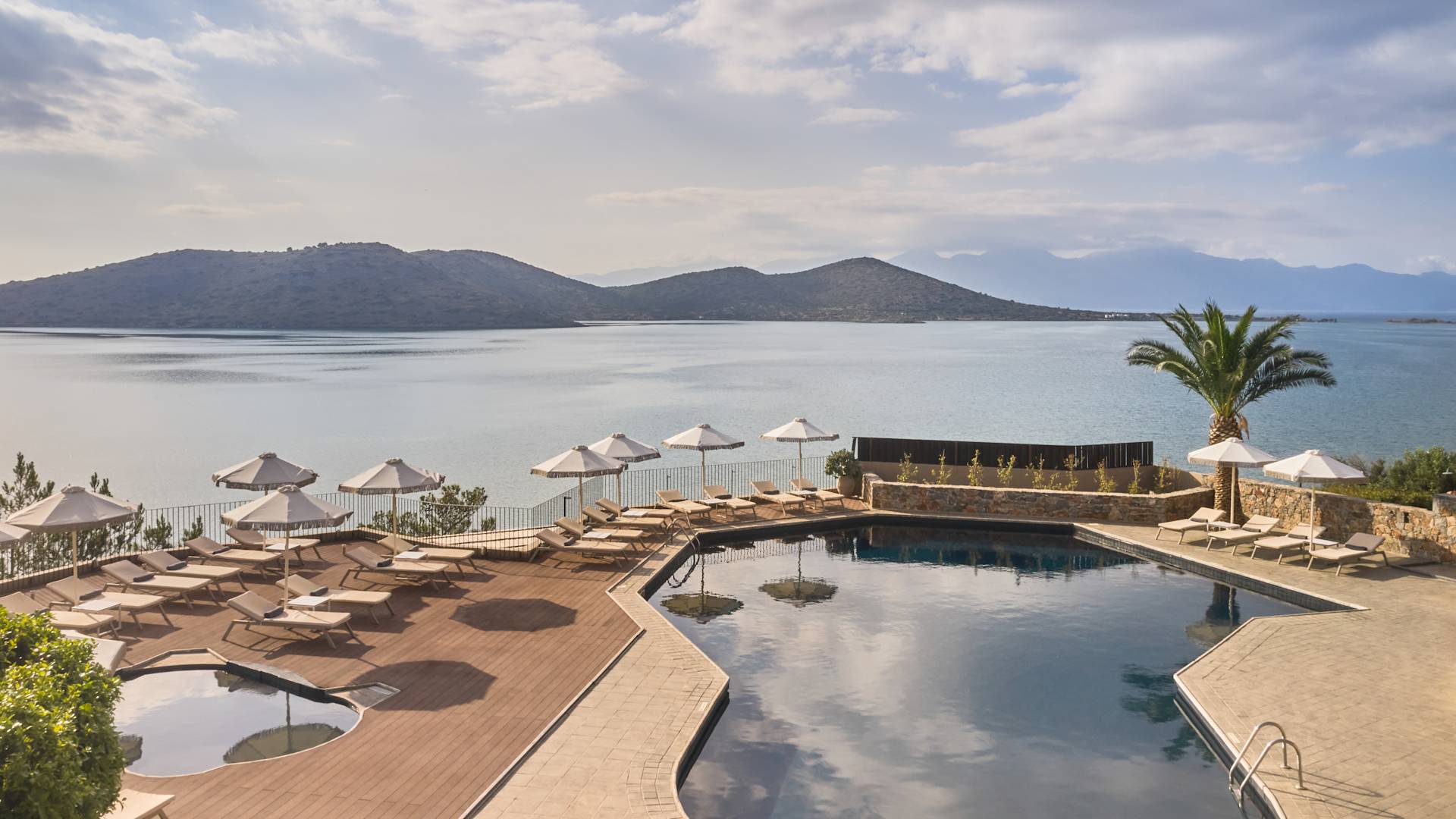 Pool area with loungers and umbrellas overlooking a calm body of water and distant mountains.