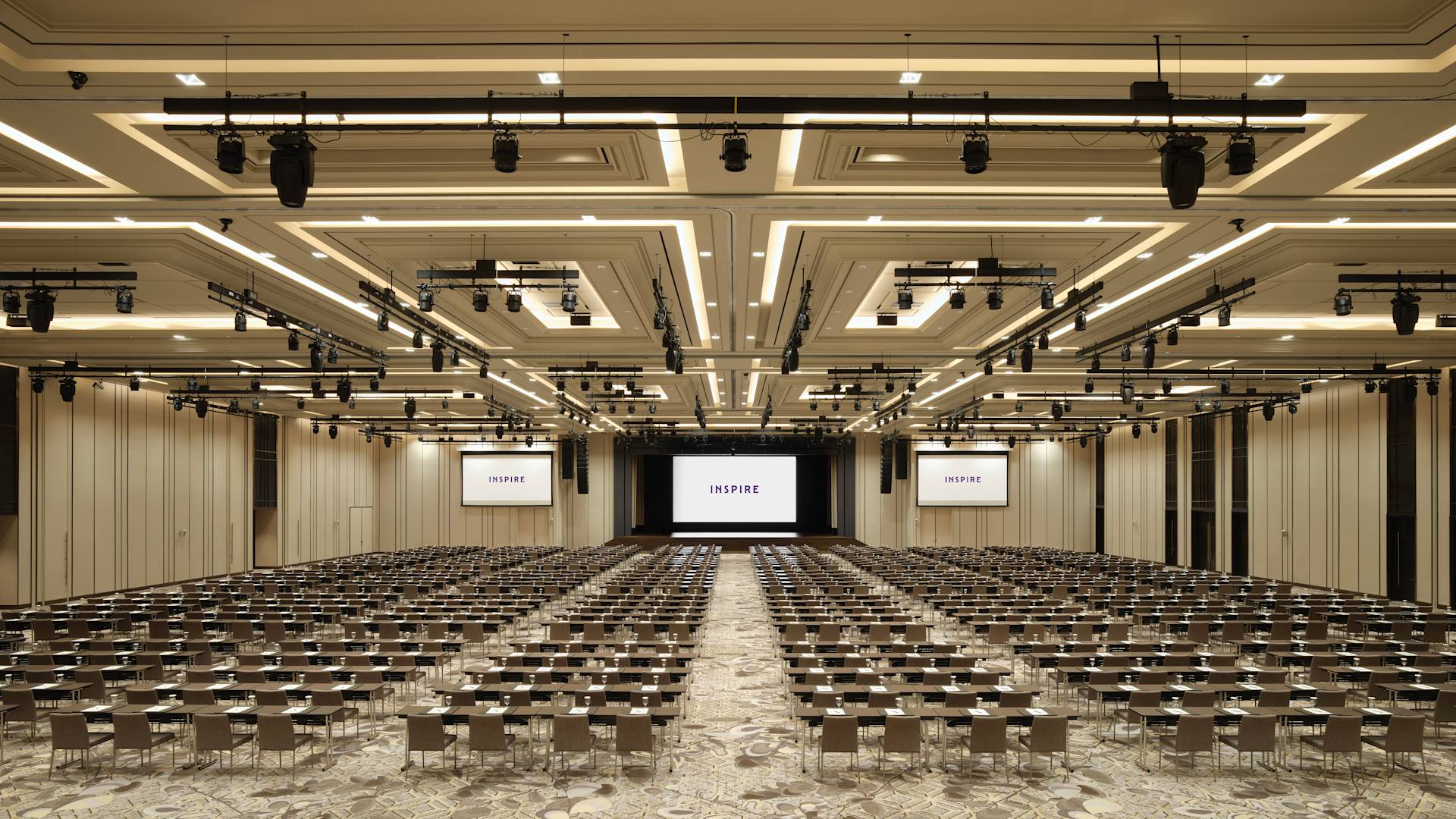 Ballroom besar dengan meja dan kursi menghadap ke beberapa layar proyektor yang menampilkan logo properti