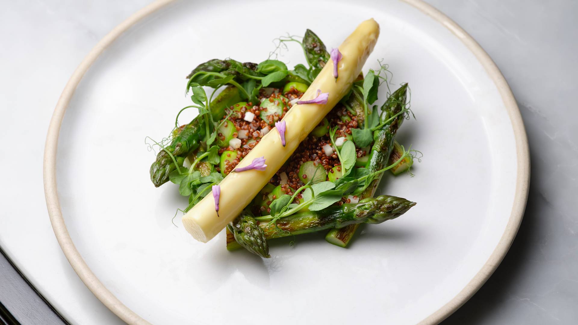 A plate with a single white asparagus spear, surrounded by green asparagus, herbs, and small edible flowers.