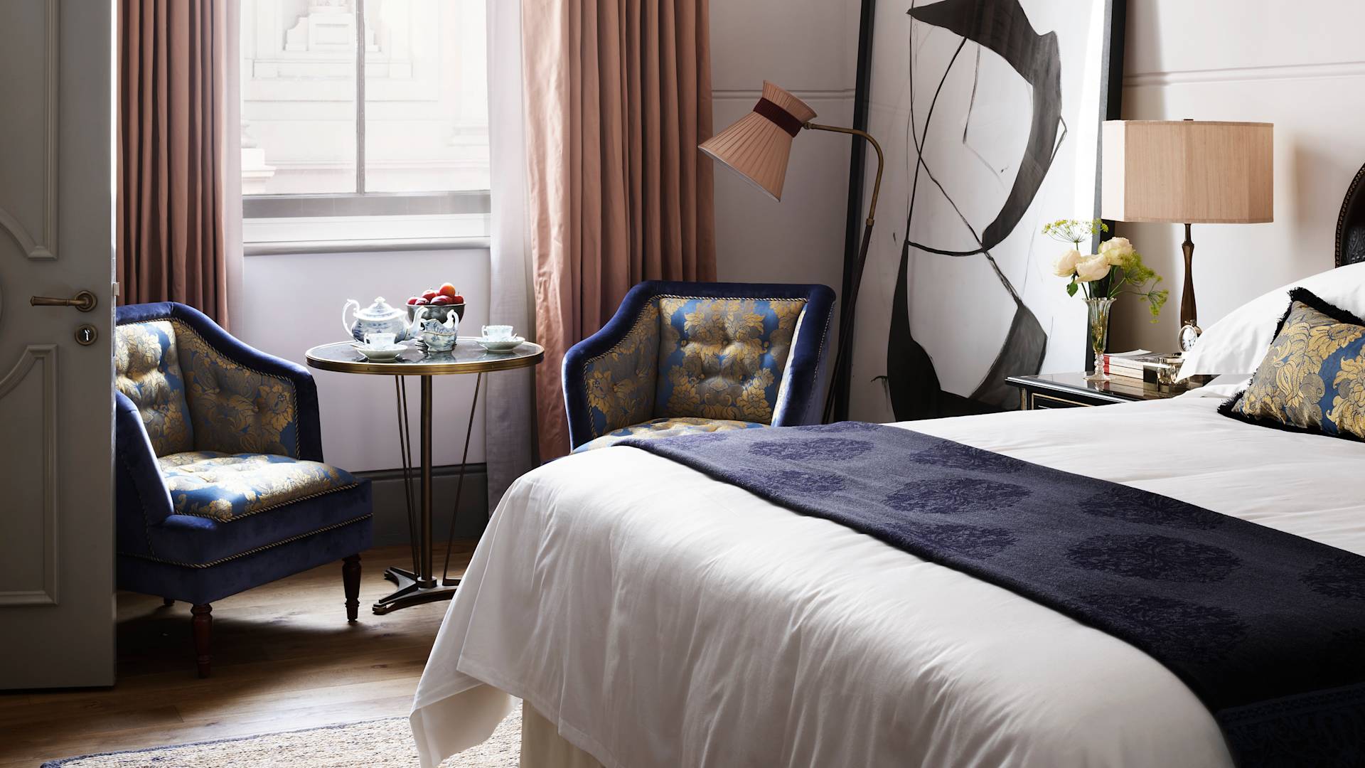 Bedroom with a large bed, blue chairs, a round table, and a large abstract painting.-next-transition