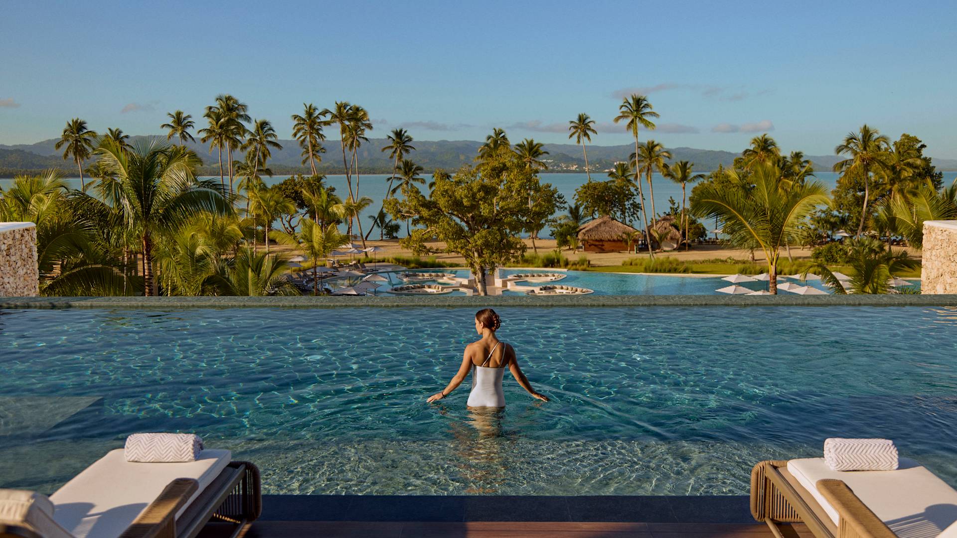 Person standing in a long infinity pool facing the ocean, surrounded by palm trees, lounge chairs, and layered pools across a tropical landscape.-previous-transition