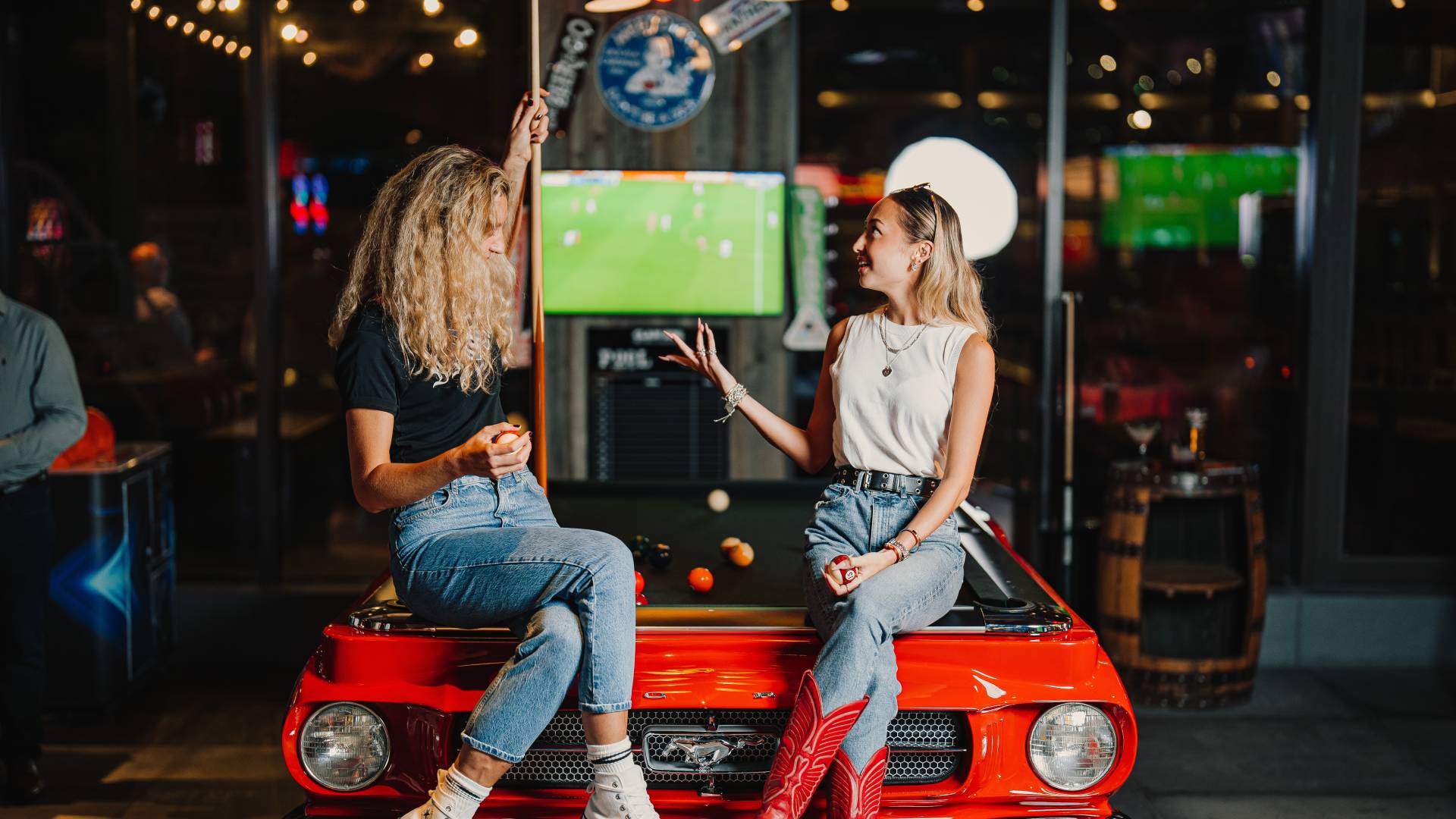 Two woman sitting on a pool table shaped like a car