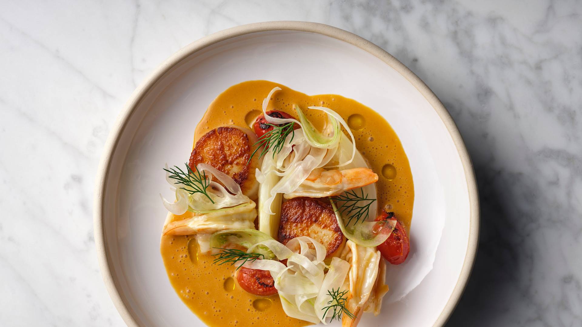 White plate with seared scallops, fennel, cherry tomatoes, and dill on a golden brown sauce, placed on a marble surface.-next-transition